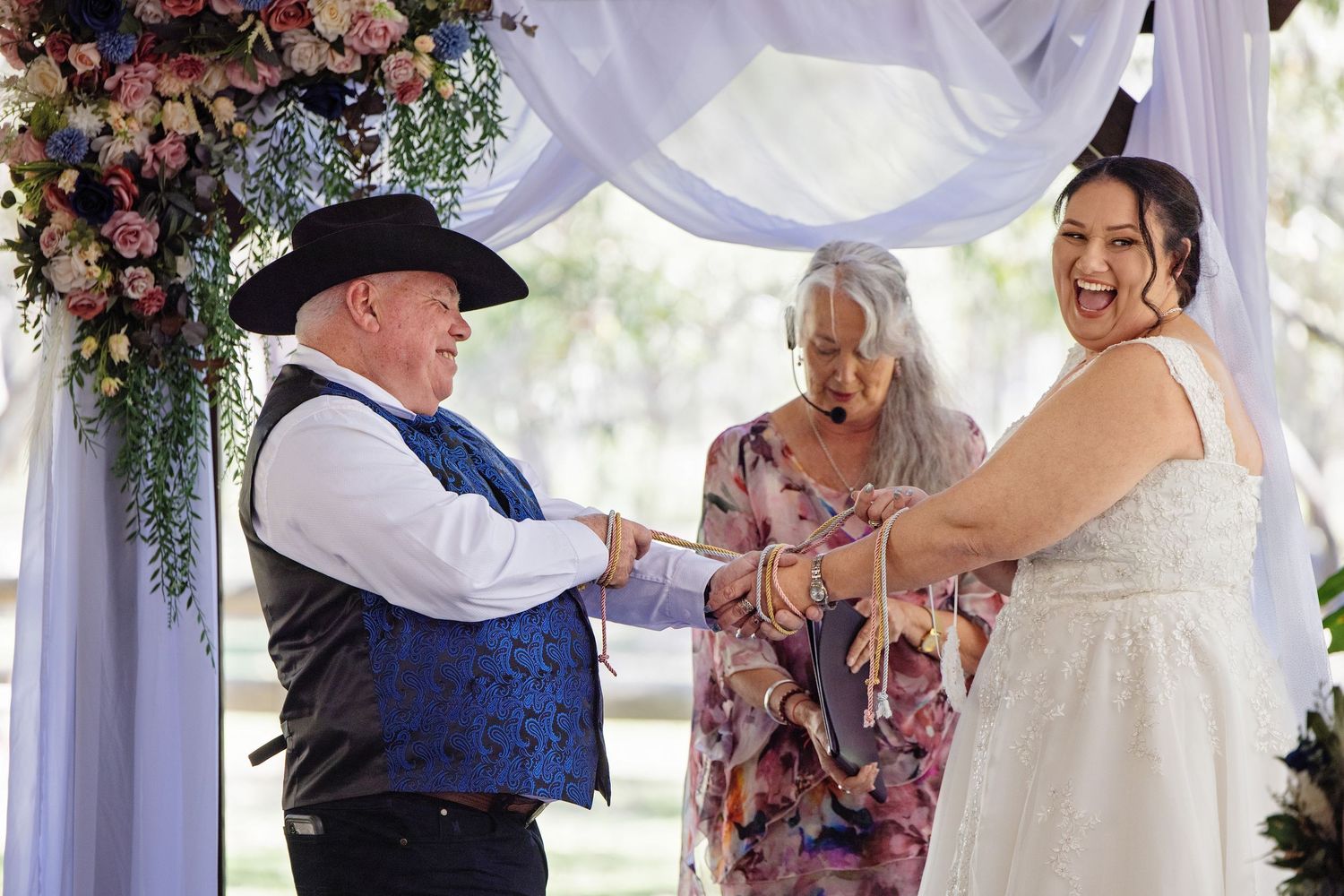 Joyful bride laughing during outdoor wedding ceremony under floral arch with groom in cowboy hat and officiant.