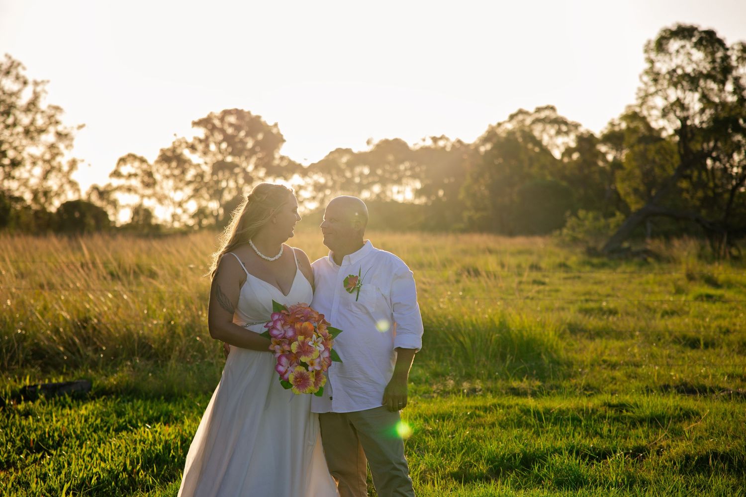 A couple in formal attire embrace in a golden sunlit field at sunset with trees in the background.