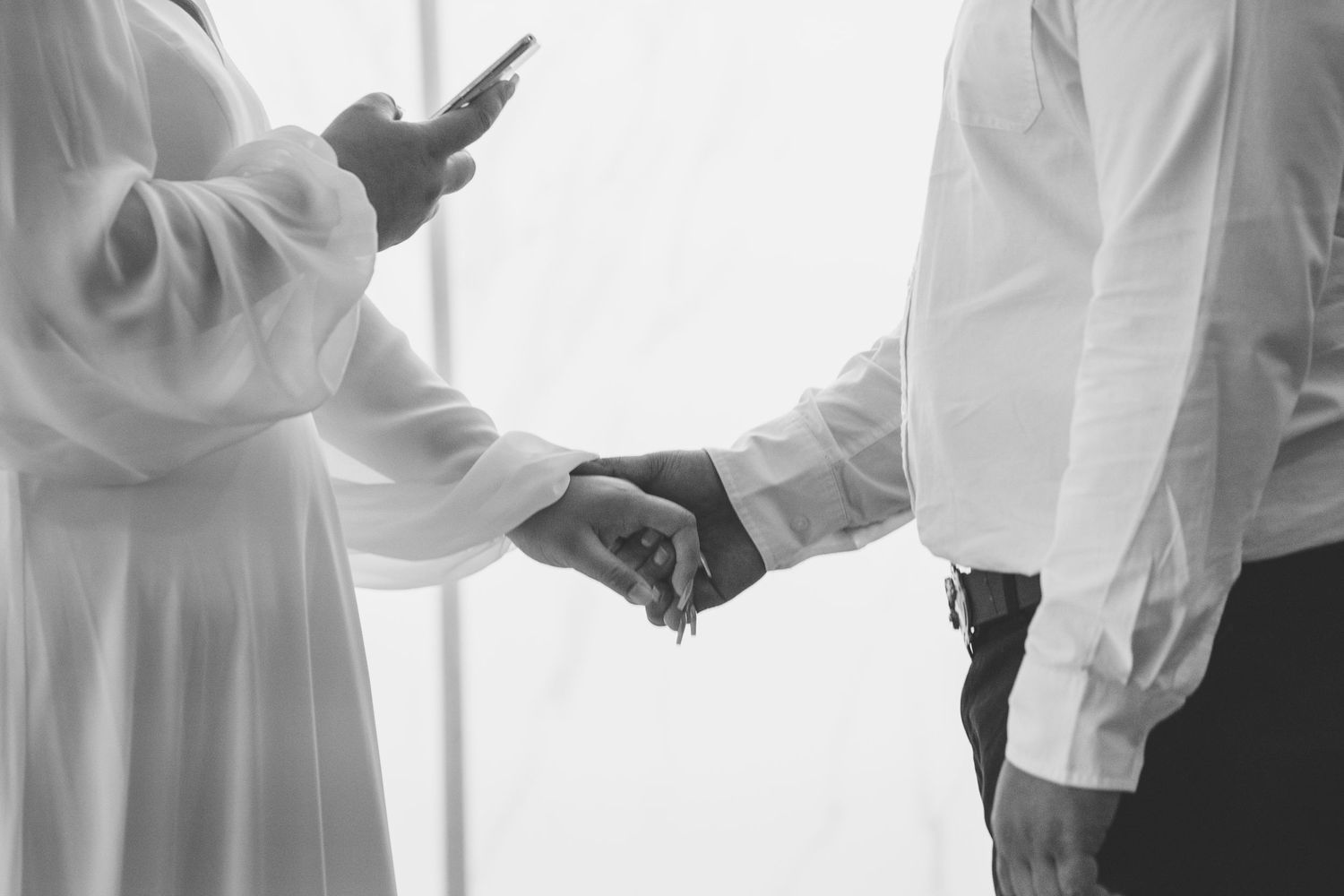 Two people in white outfits hold hands while one checks a phone in this black and white romantic moment.
