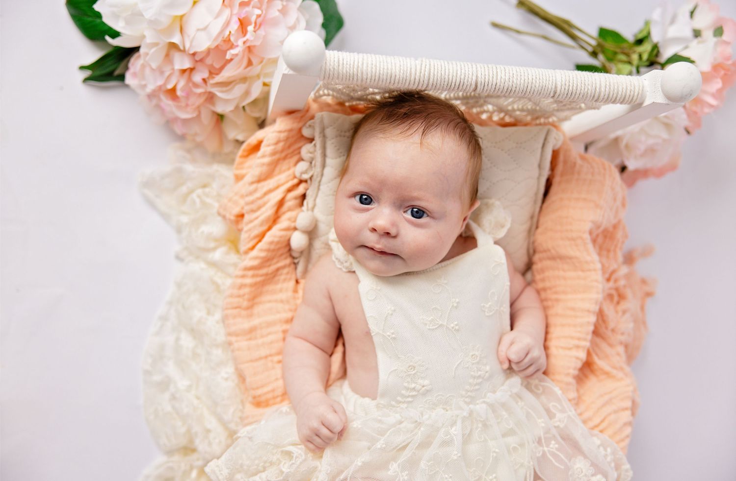A baby in a white outfit lies on a cream-colored blanket with peach and green floral decorations.