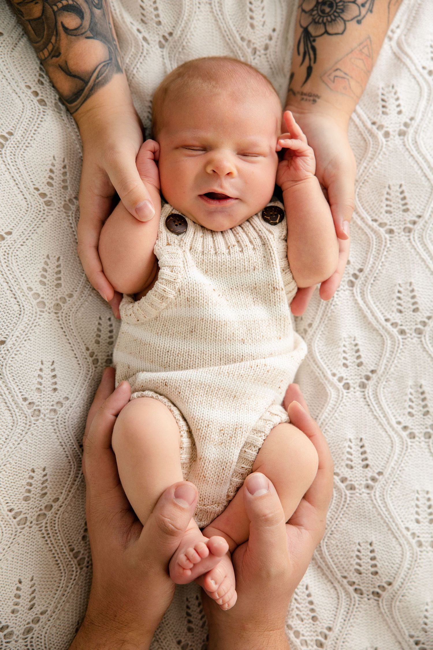 Newborn baby in knit romper held gently by tattooed parents hands on white blanket.