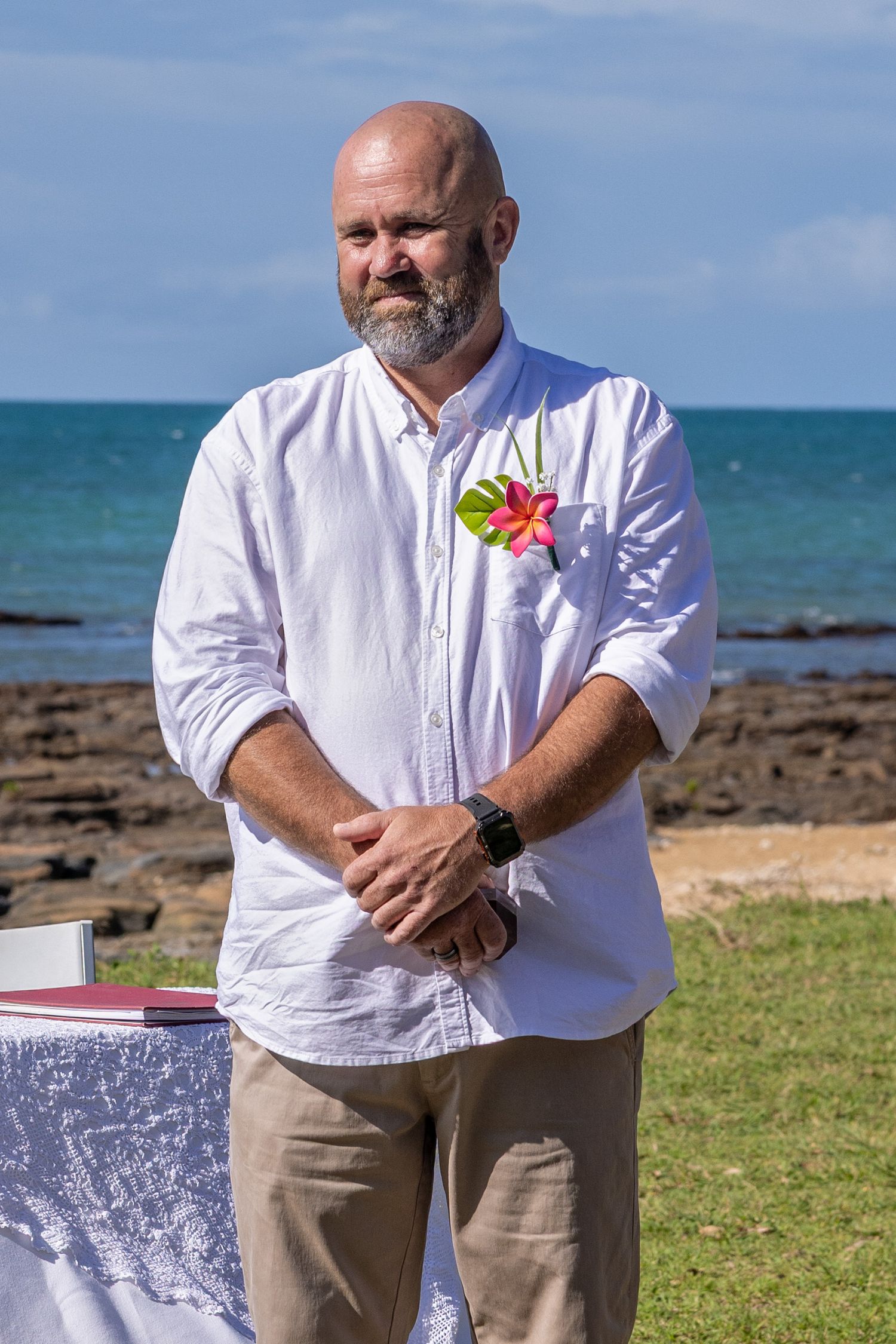 Someone wearing a white button-down shirt with a tropical flower stands by the ocean at a beach event.