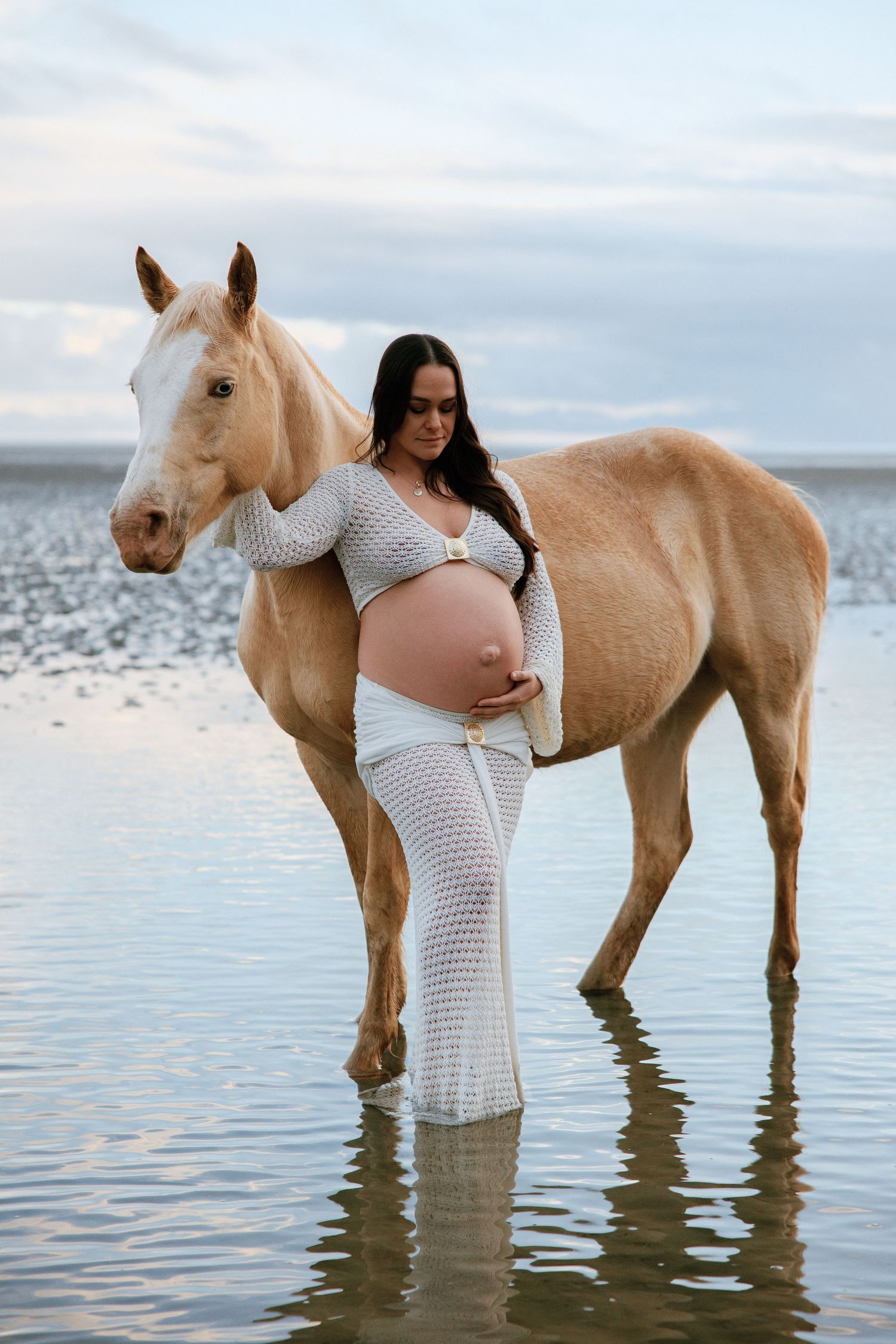 Pregnant woman in white crochet dress stands beside a palomino horse in shallow water at sunset.