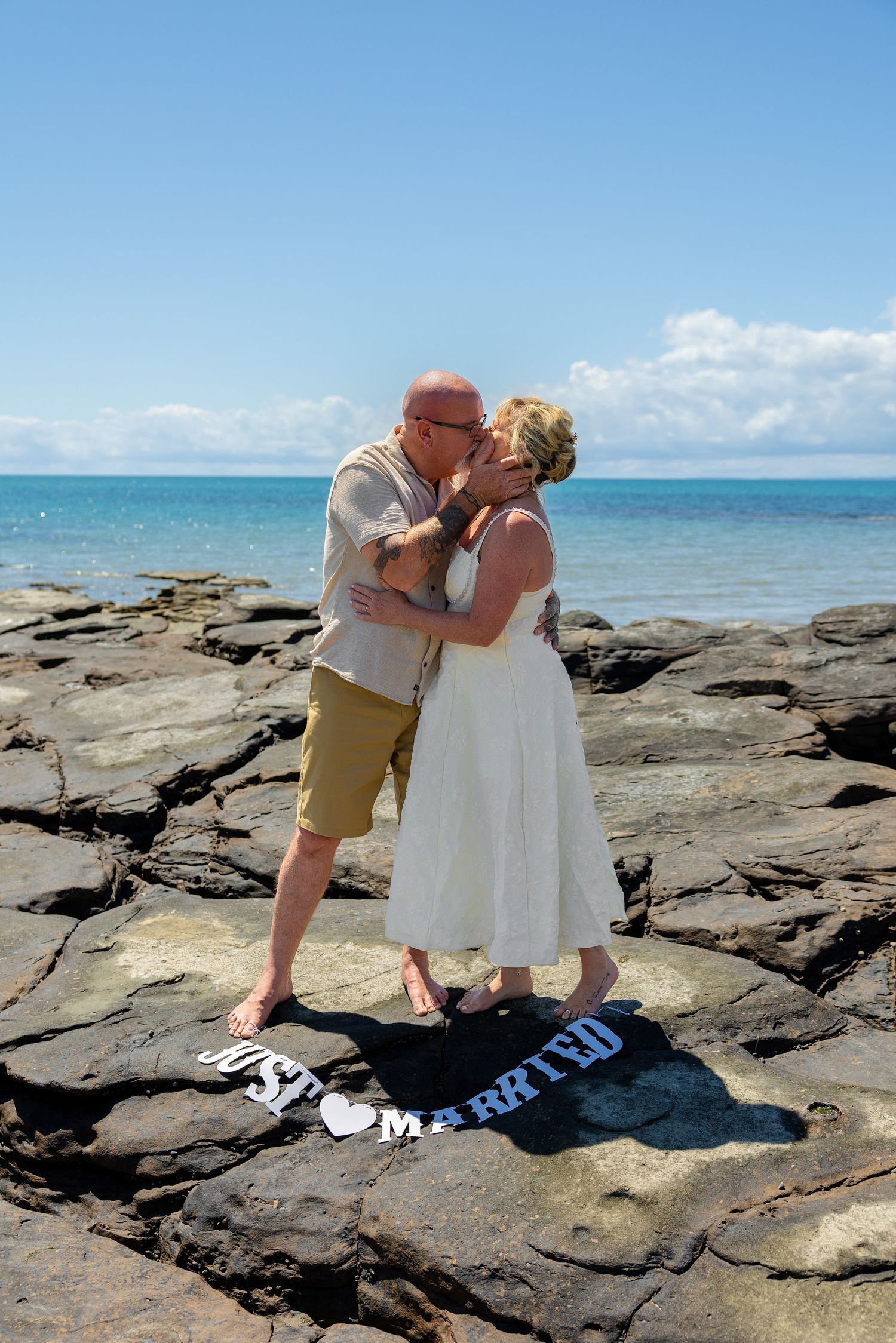 Newlywed couple kissing on rocky beach with ocean backdrop, just married sign at their feet on sunny day.