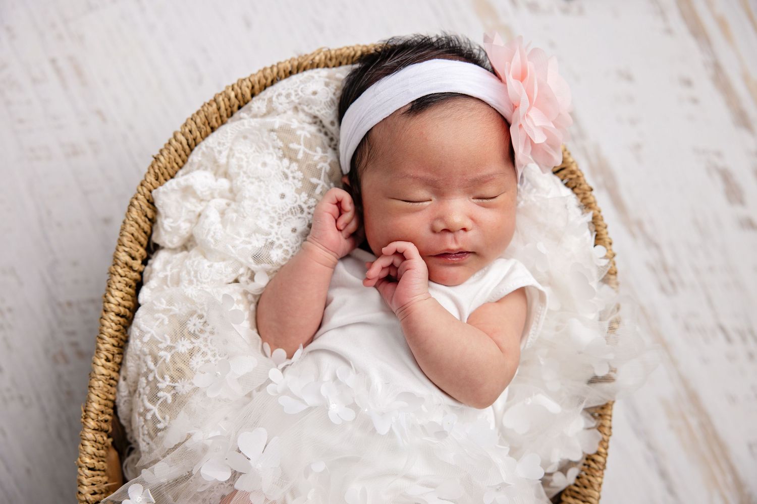 Newborn baby sleeping peacefully in a woven basket surrounded by delicate white lace fabric.