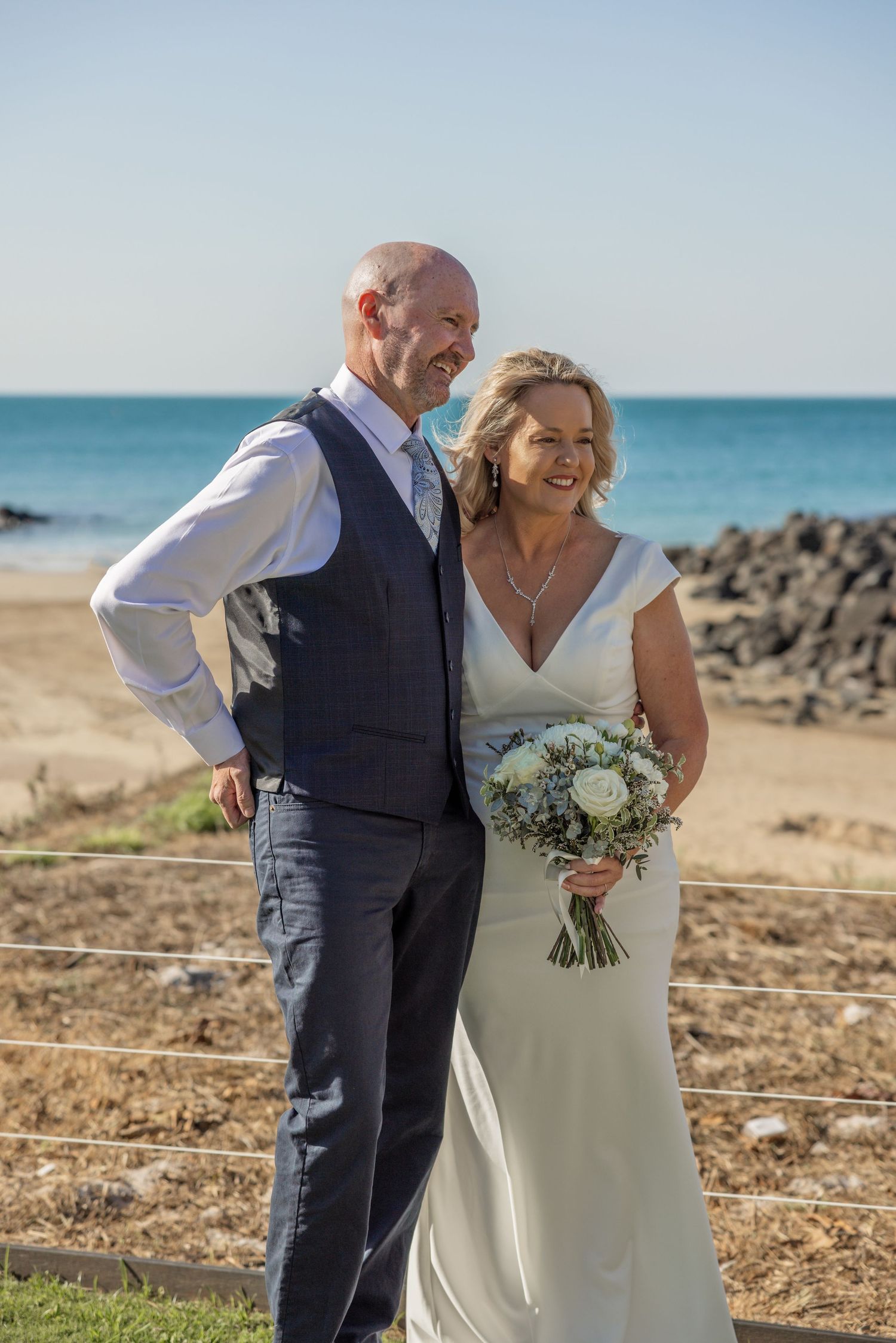 A couple shares a romantic moment on a sunny beach with rocky shoreline and blue ocean in the background.
