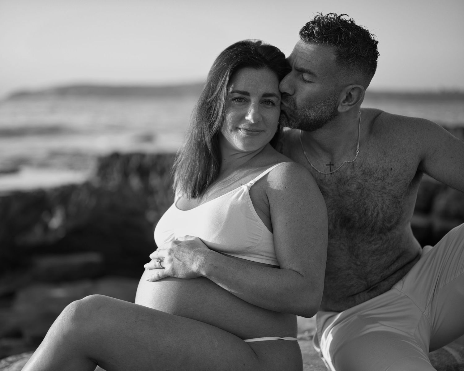 A romantic black and white maternity photo shoot on a beach with a couple embracing.