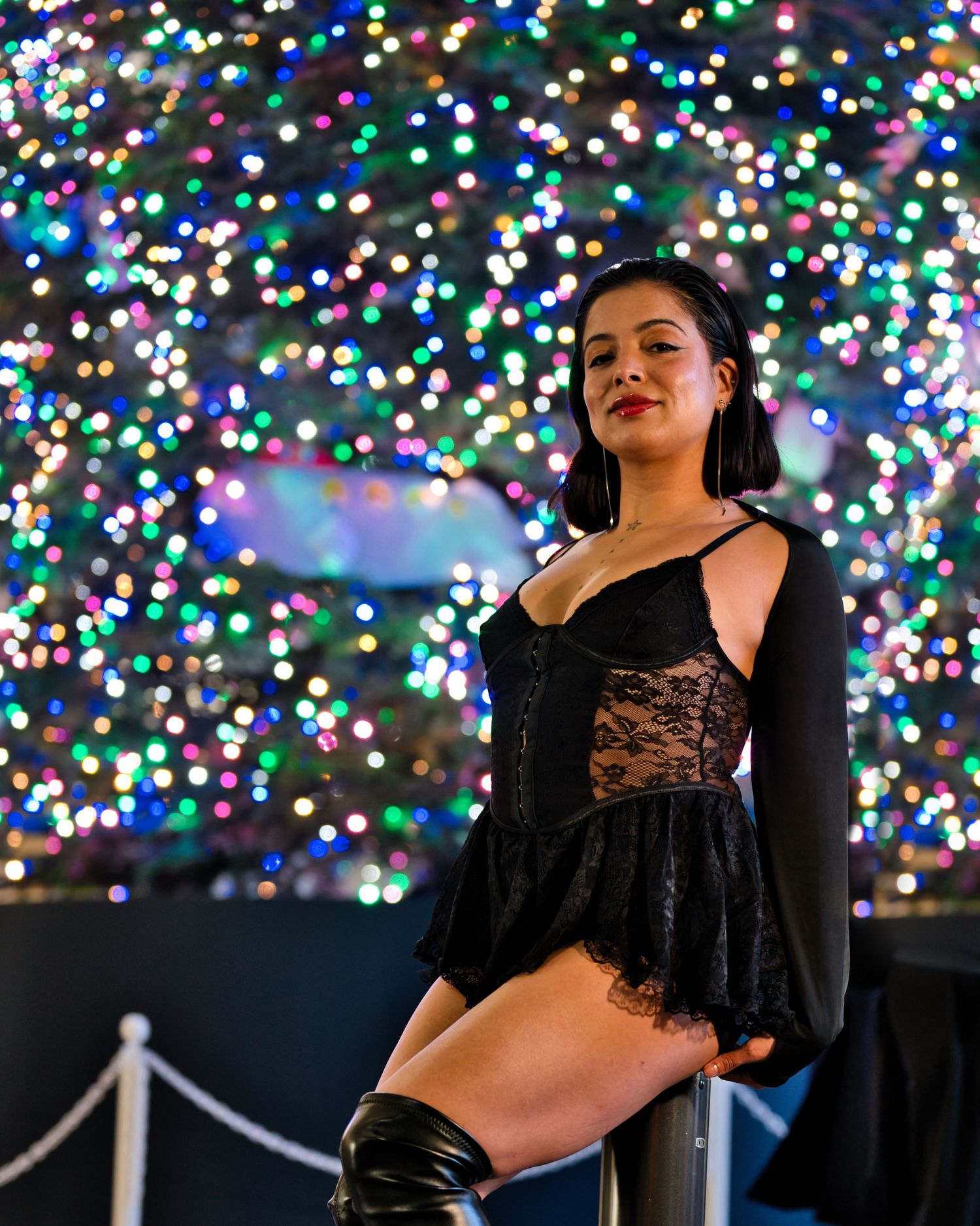 Stylish figure poses against backdrop of colorful holiday lights display.