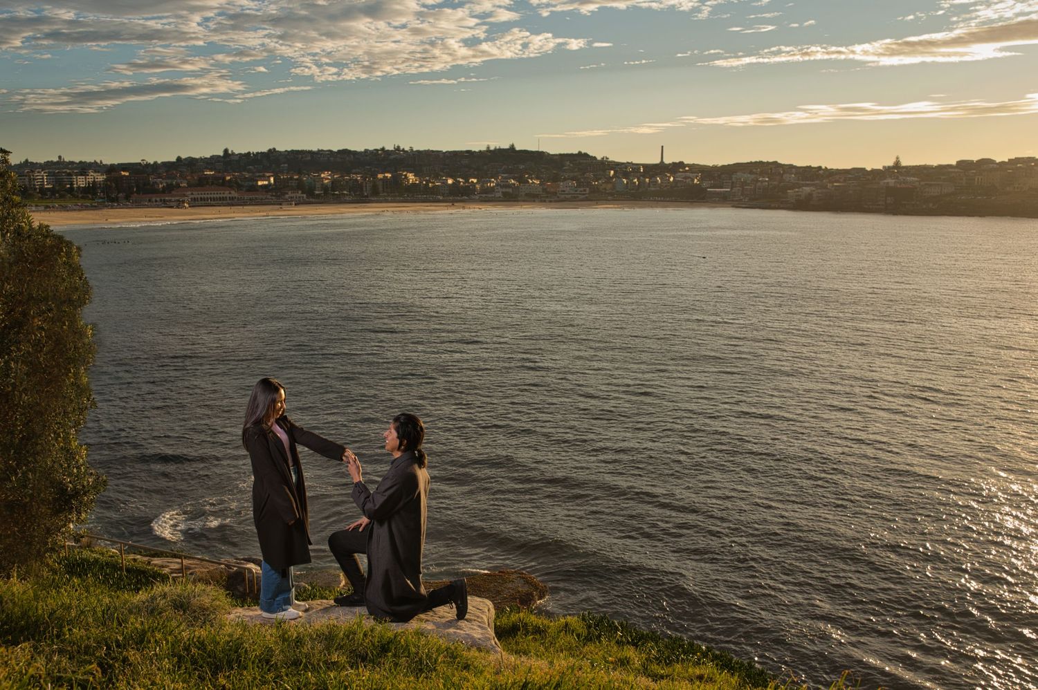 A romantic sunset proposal scene on a grassy cliff overlooking a waterfront with beautiful golden sky.