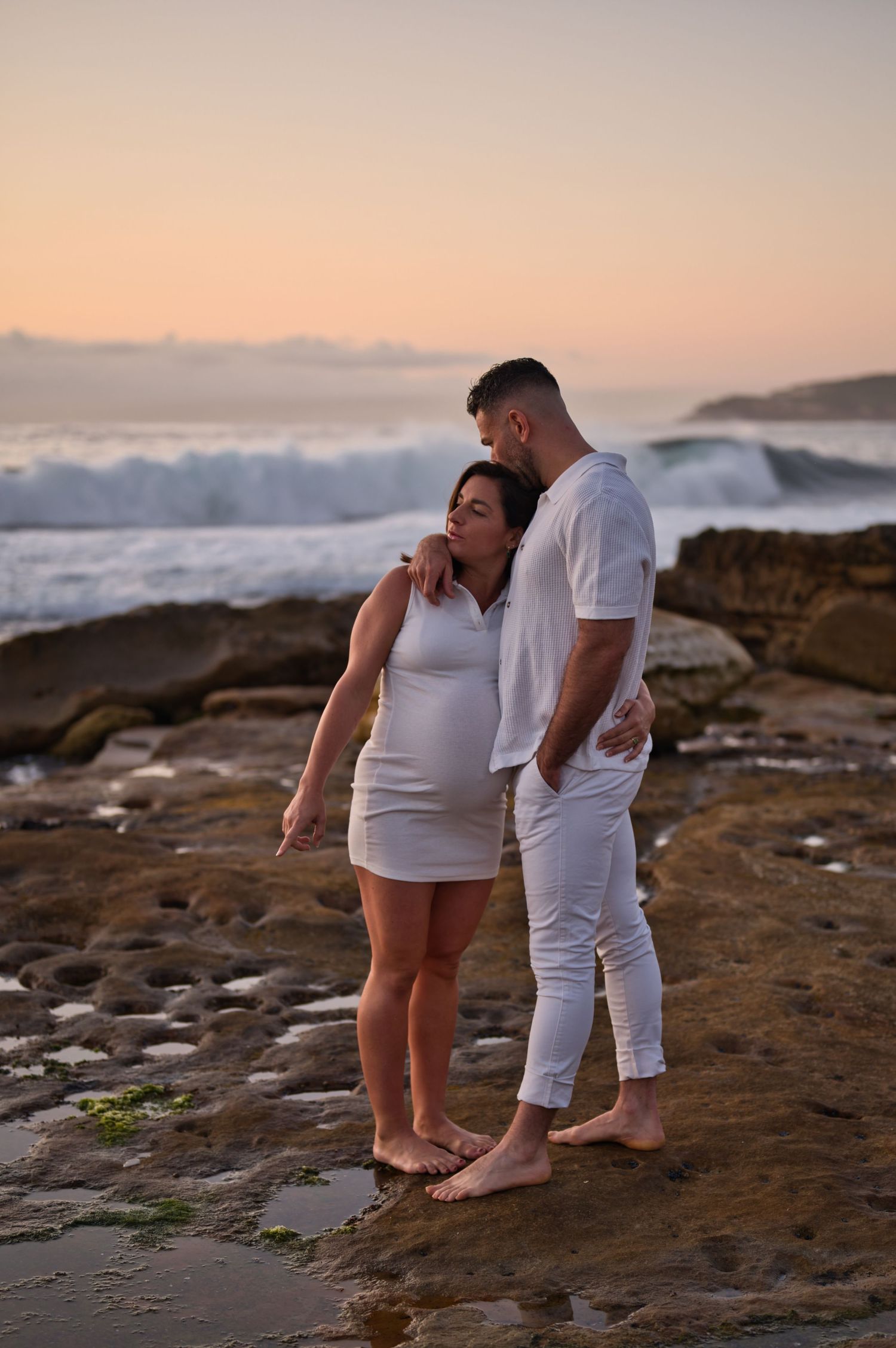 A couple embraces on a rocky beach at sunset with crashing waves in the background.