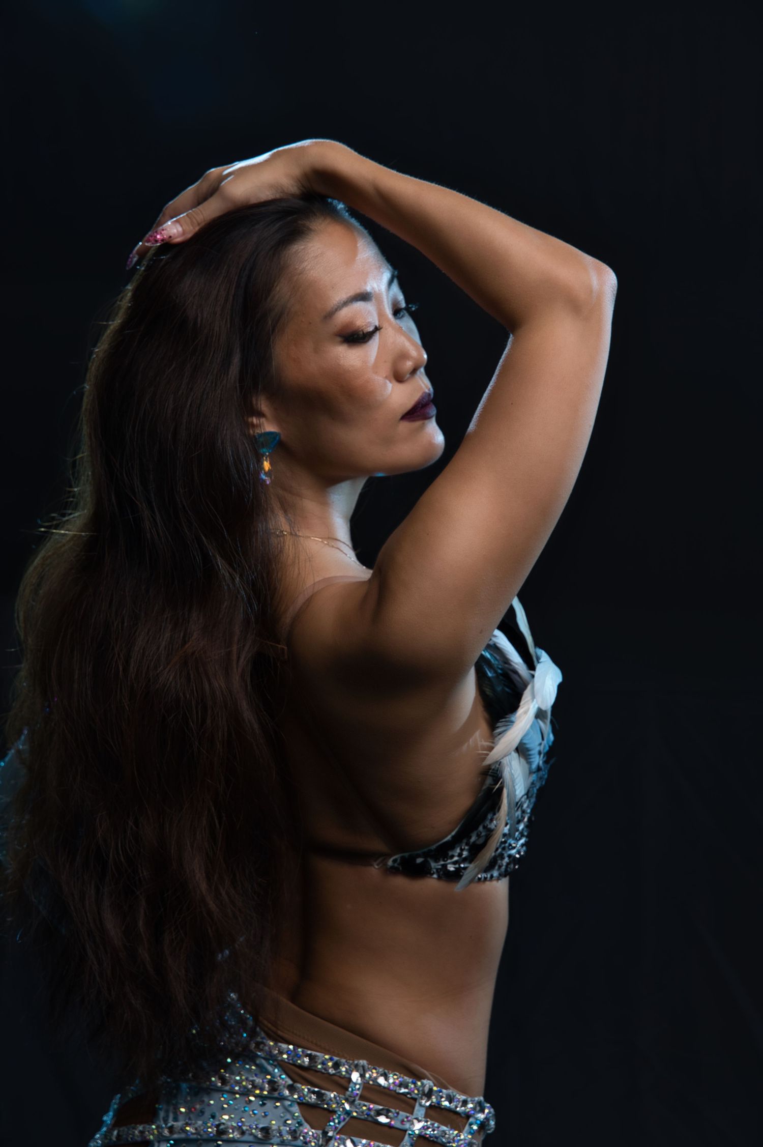 Dancer wearing blue sequined costume poses elegantly with raised arms in moody lighting.