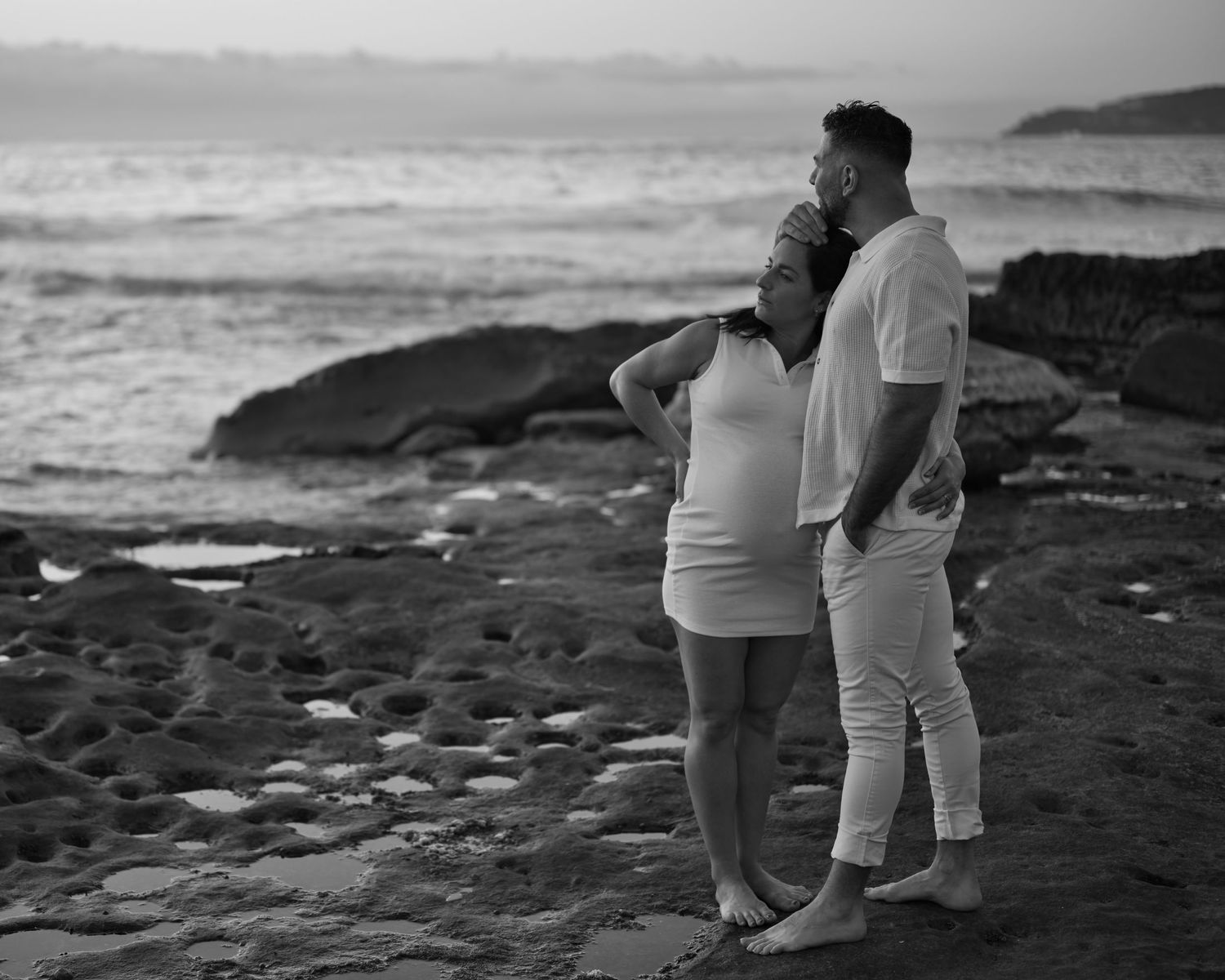 Silhouetted couple stands together on rocky beach at sunset.