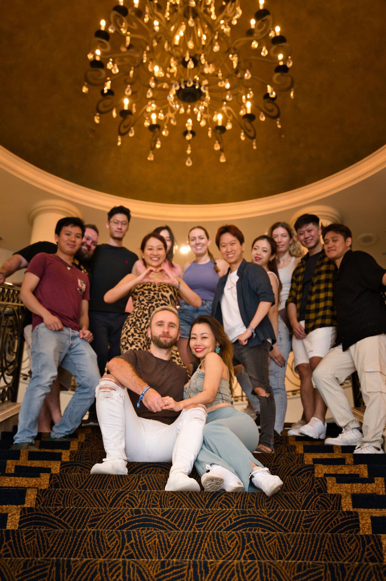 Group photo of dance performers beneath an ornate chandelier on carpeted steps.
