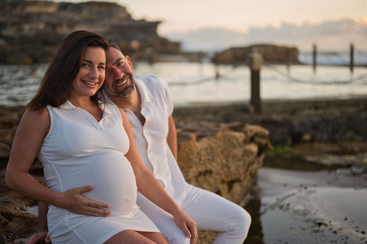 A couple in white clothing poses together near the ocean at sunset.
