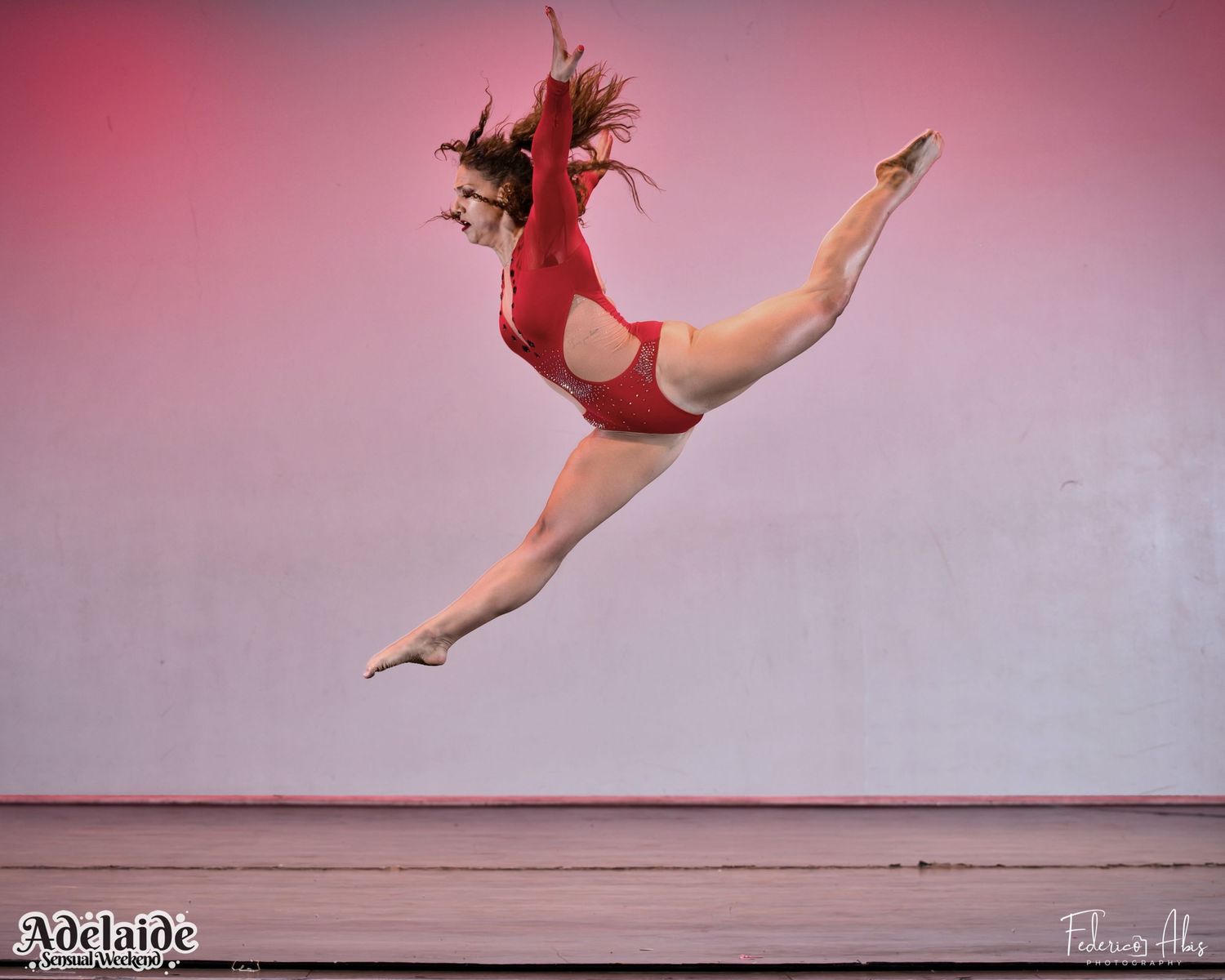 Dancer in red costume performing mid-air split leap against pink backdrop.