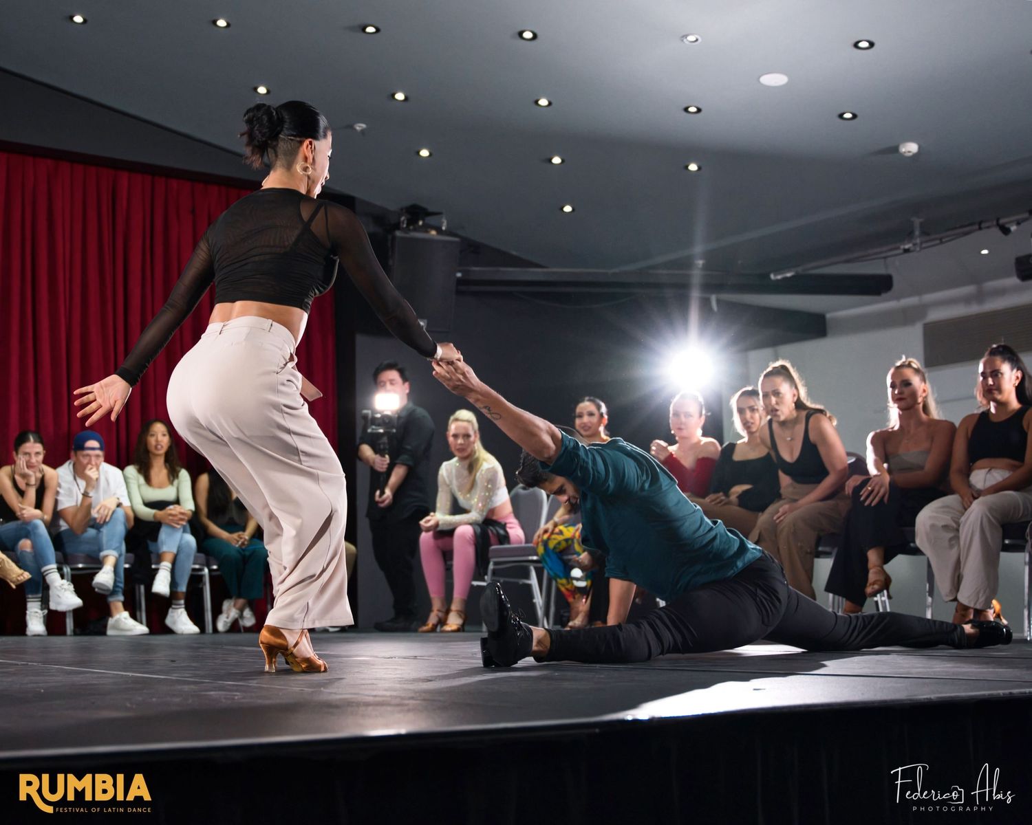 Dancers perform a dramatic dip move on stage in front of an audience with red curtains.