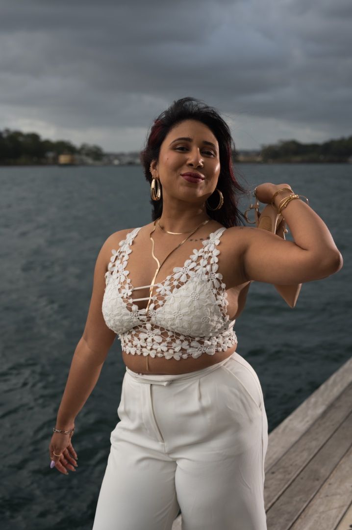 A woman wearing a white lace top and pants poses on a wooden pier overlooking the water.