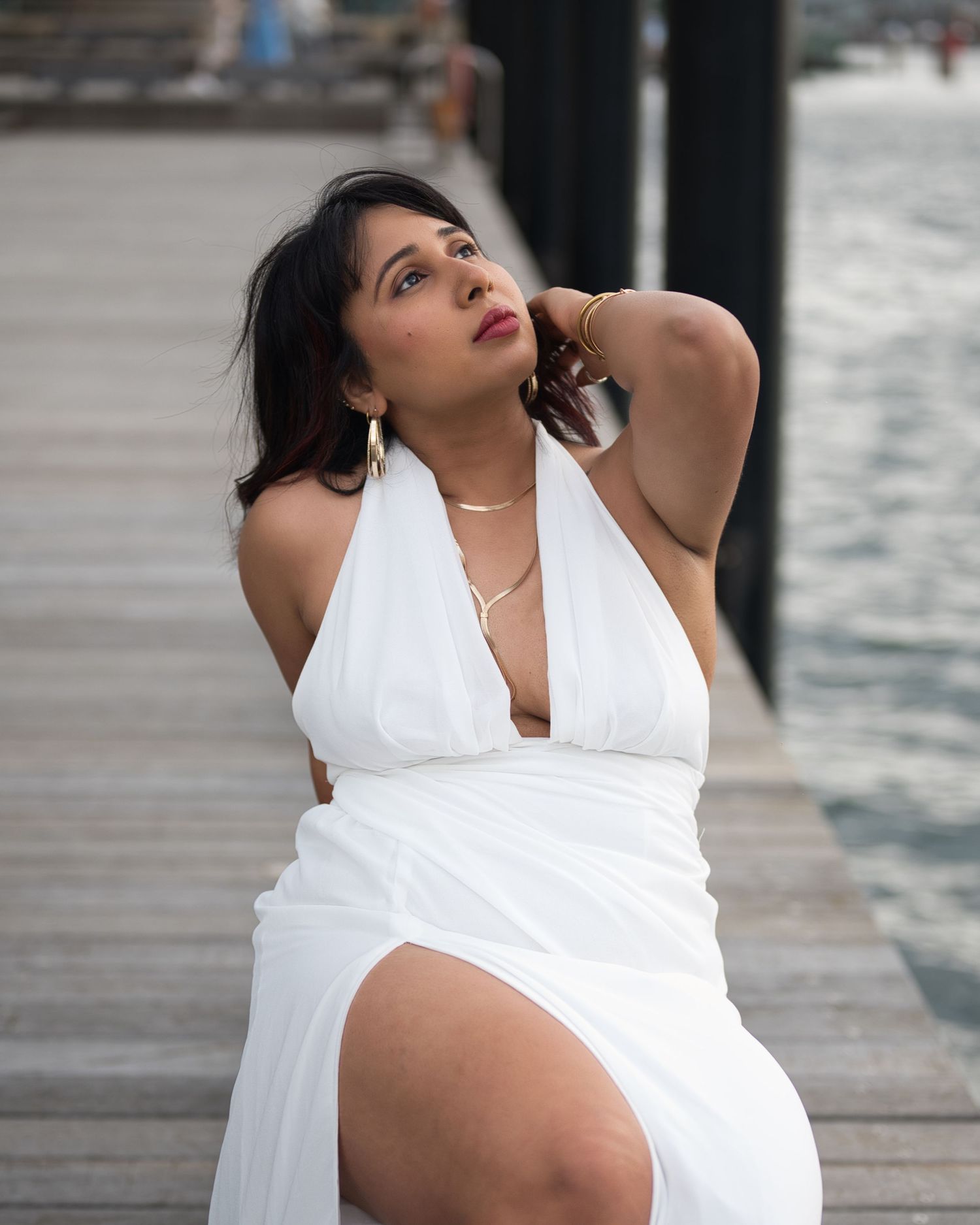 A woman in a white halter dress poses confidently on a wooden pier by the water.