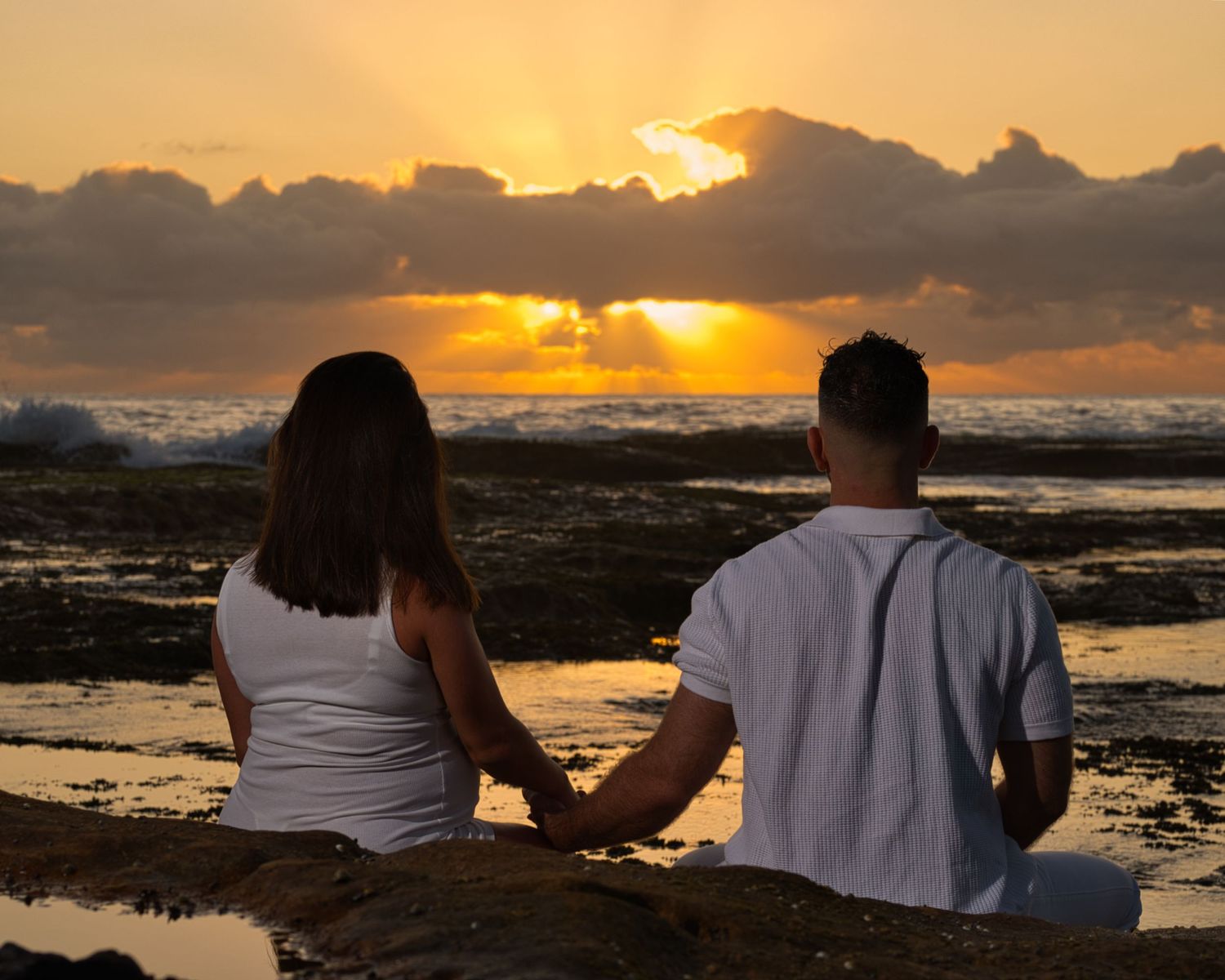 A couple holds hands while watching sunset over rocky coastal landscape.