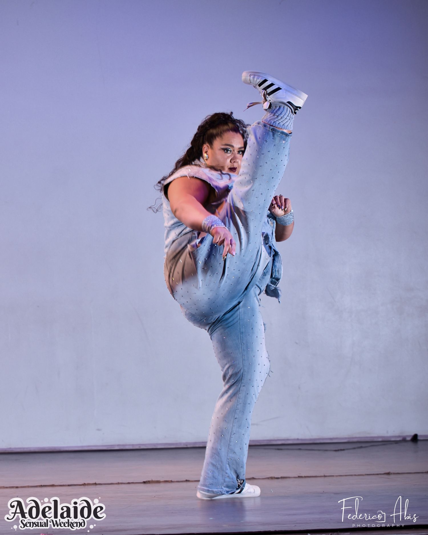 Performer executing high kick dance move in denim outfit against purple backdrop.