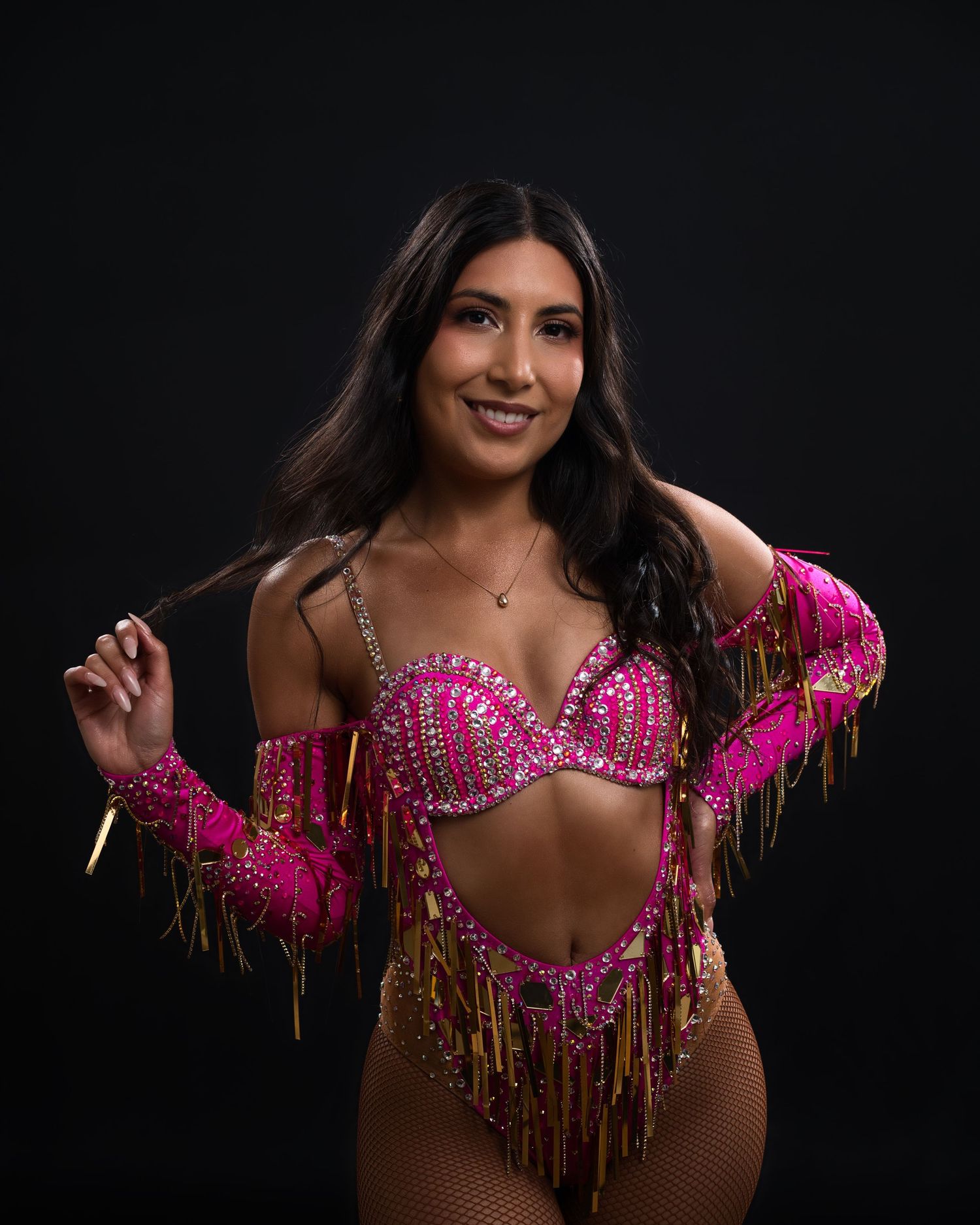 Dancer poses in vibrant pink and gold fringe bodysuit against black background.