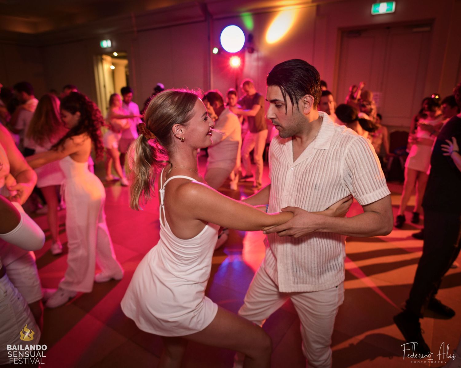 Dance partners in white outfits moving together on a dance floor with pink lighting.