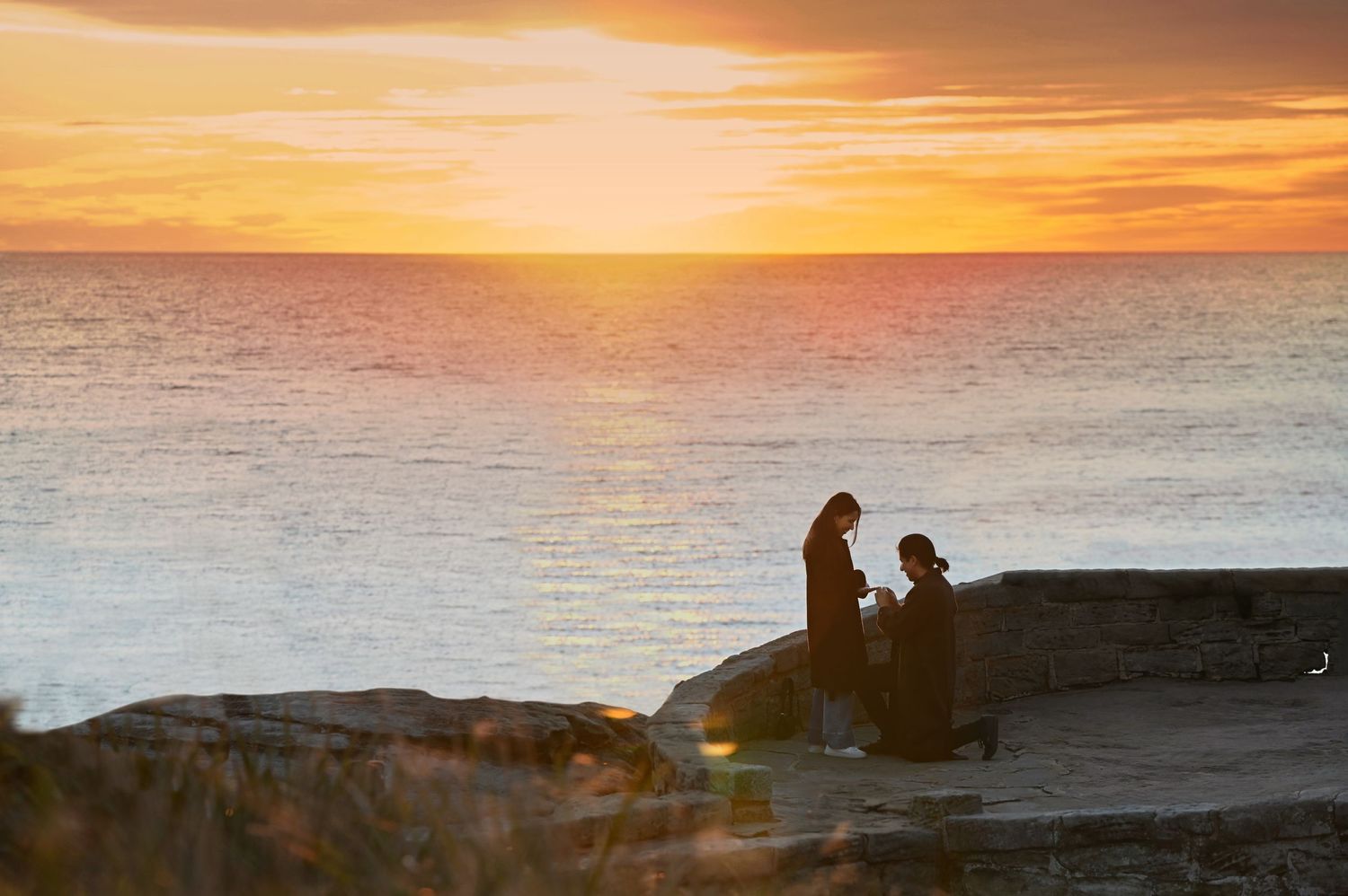Silhouettes of couple sitting on rocky coast during golden sunset over ocean.