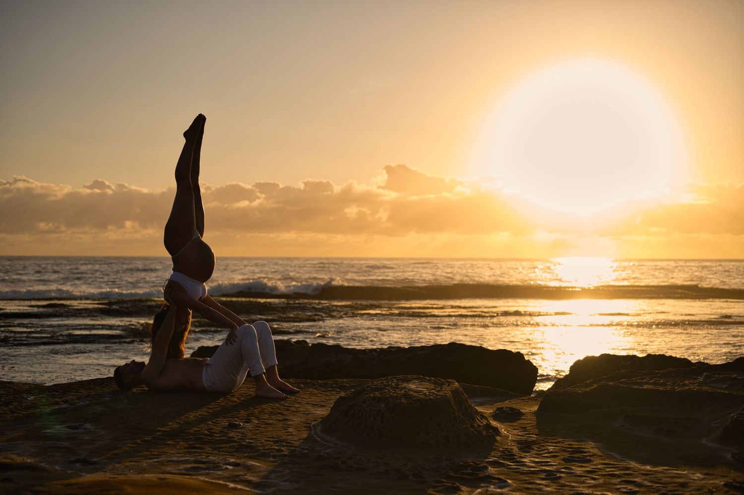 A person performs a headstand yoga pose on rocks by the ocean at sunset.