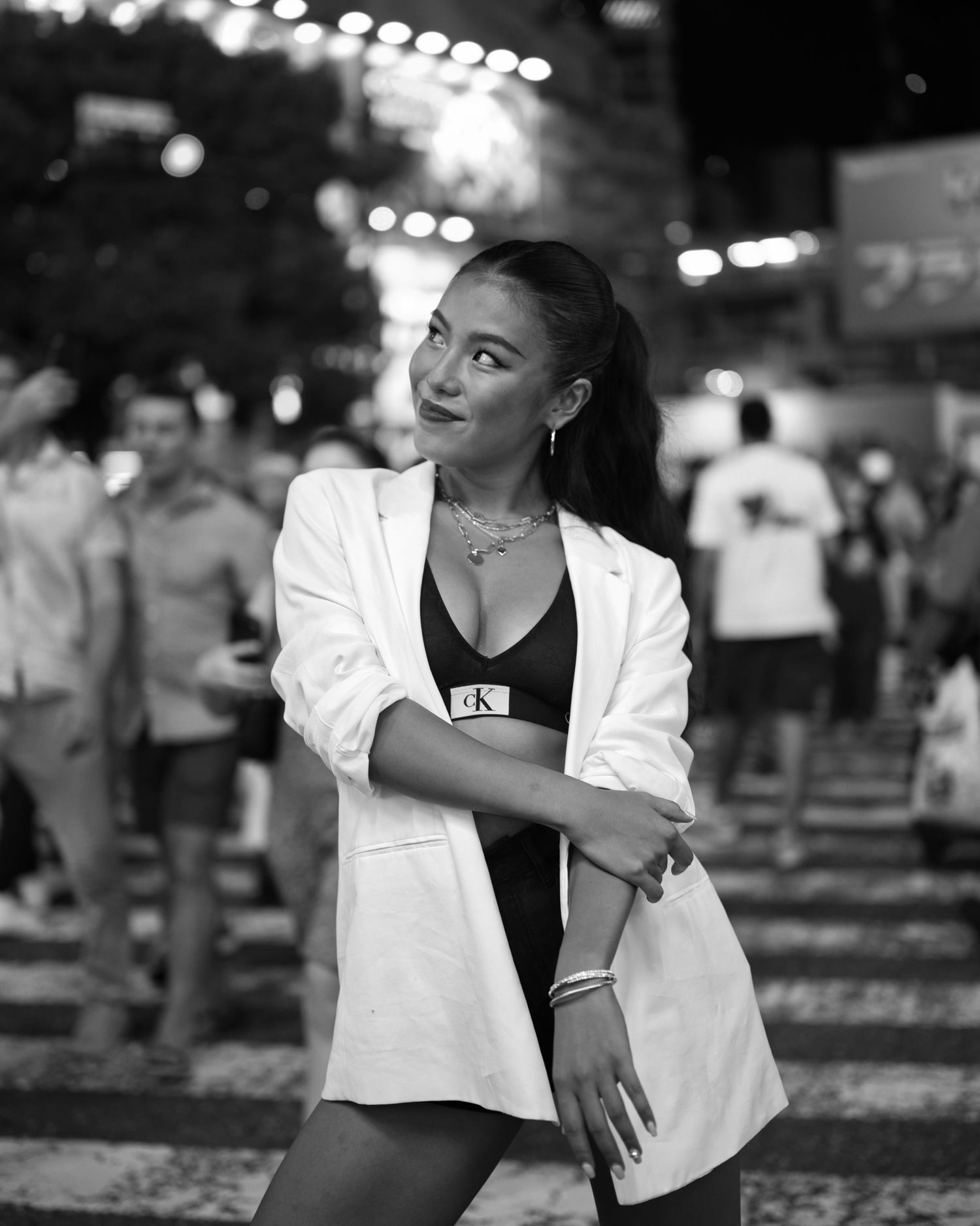 A black and white street fashion portrait with city lights and crowd in background.