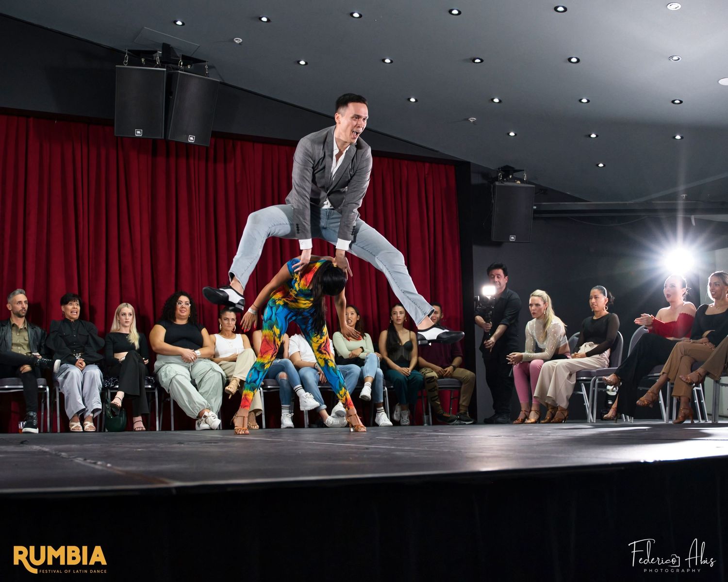 Solo dancer performs an energetic jump move on stage with audience in background.