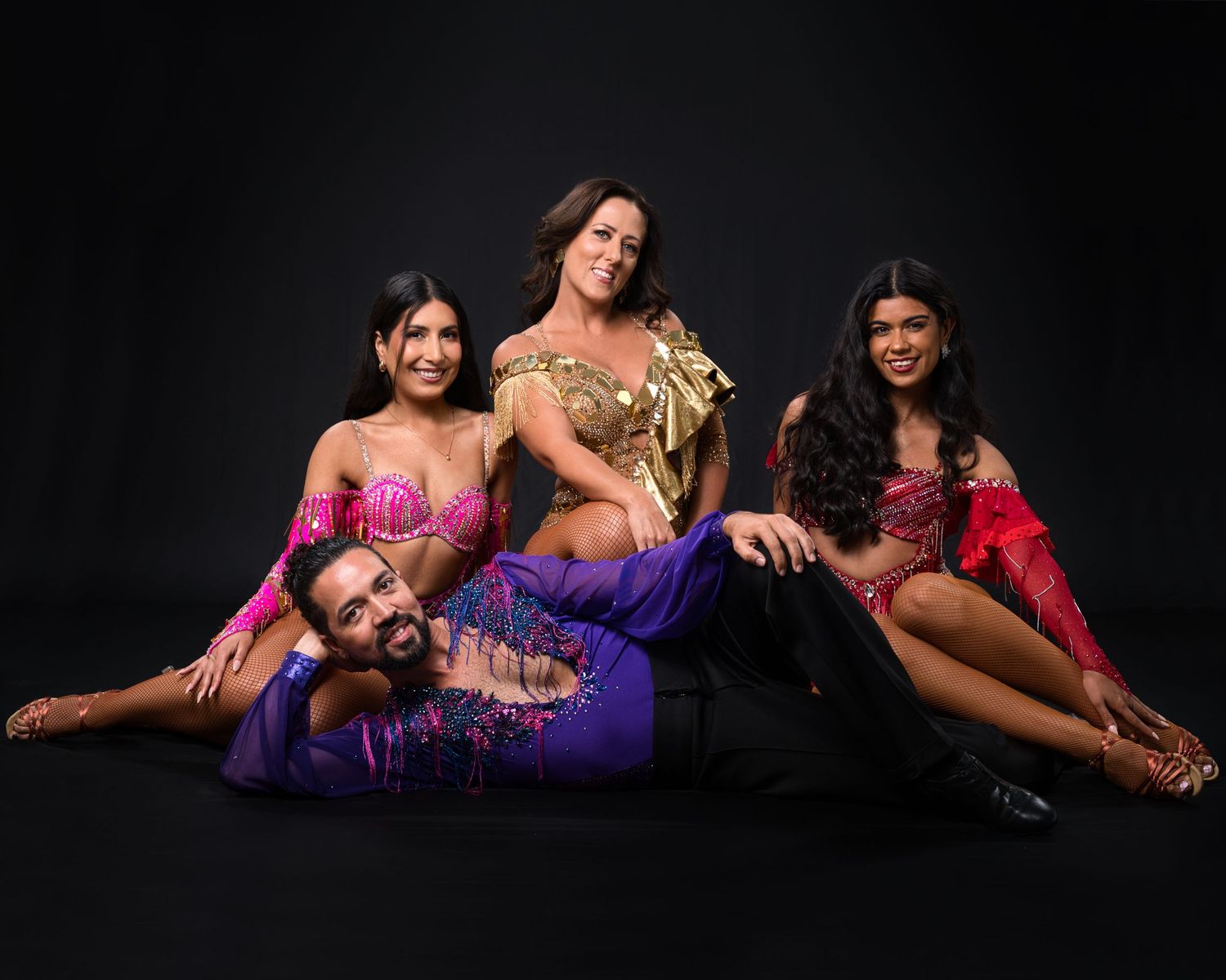 Group of dancers in colorful performance costumes posing together against black background.