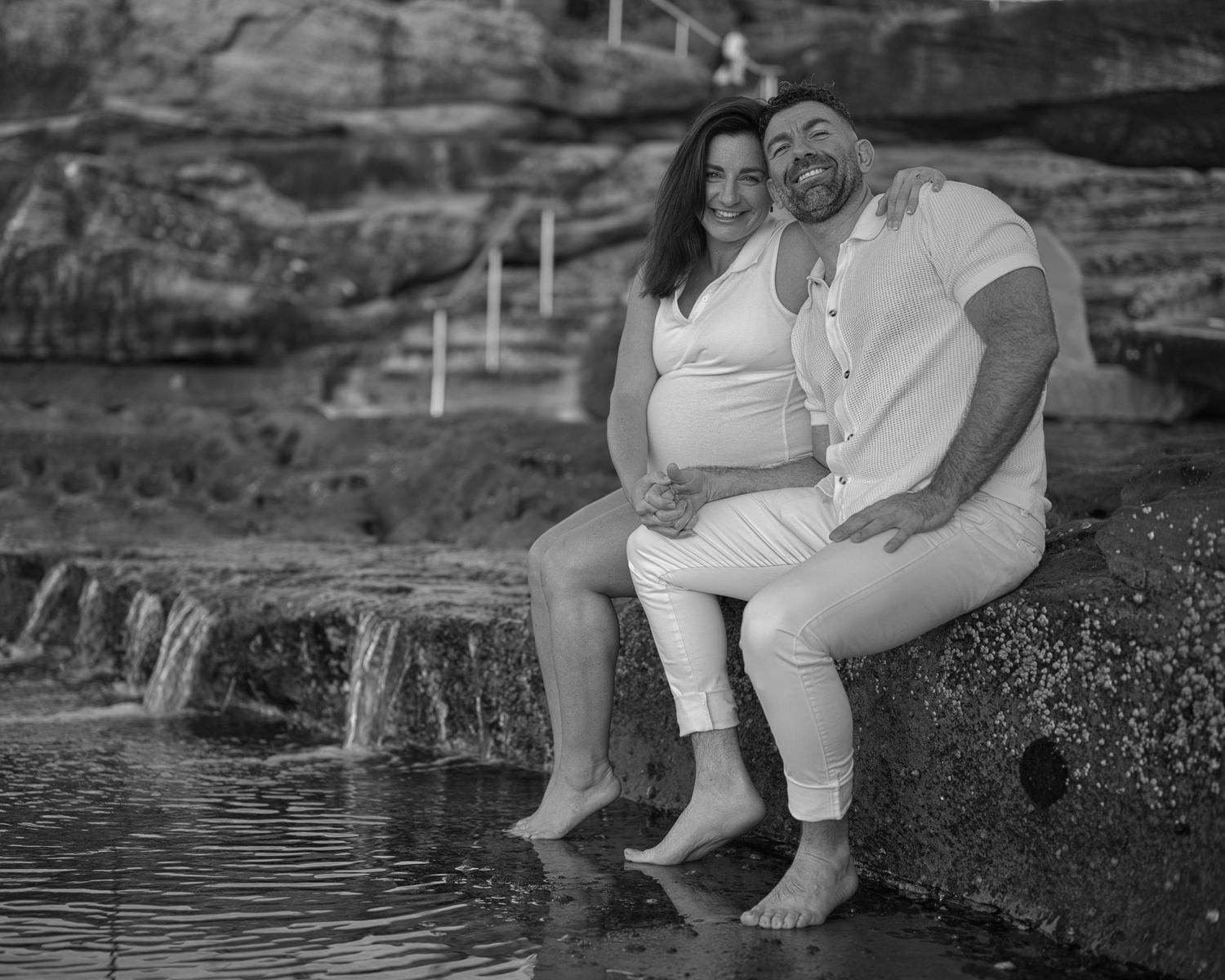 A couple sits together with their feet in a small waterfall, captured in black and white.