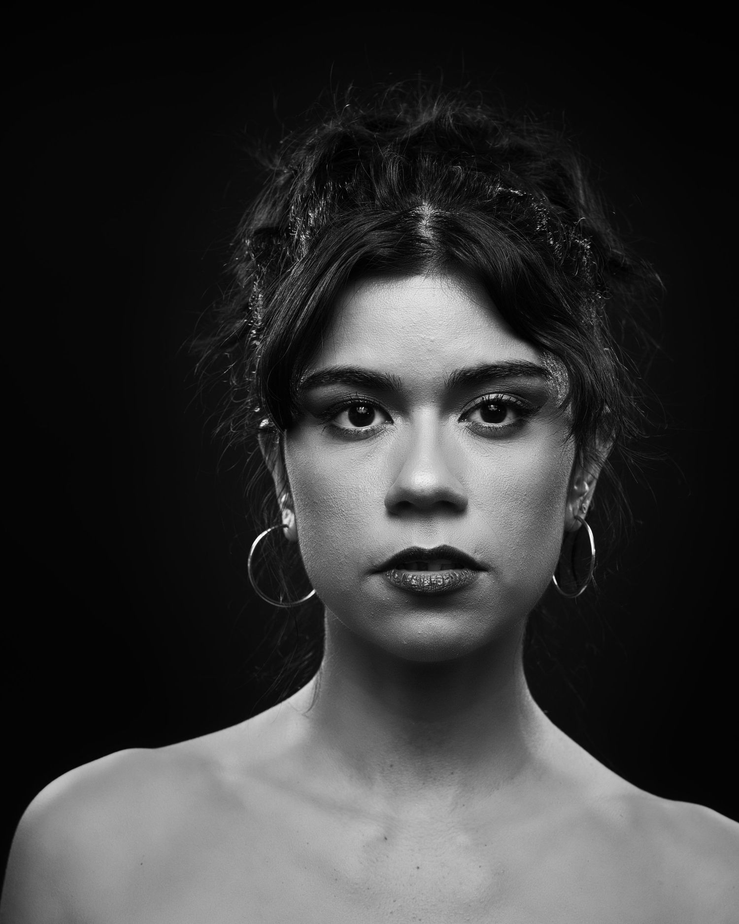 Dramatic black and white portrait with silver hoop earrings against dark background.