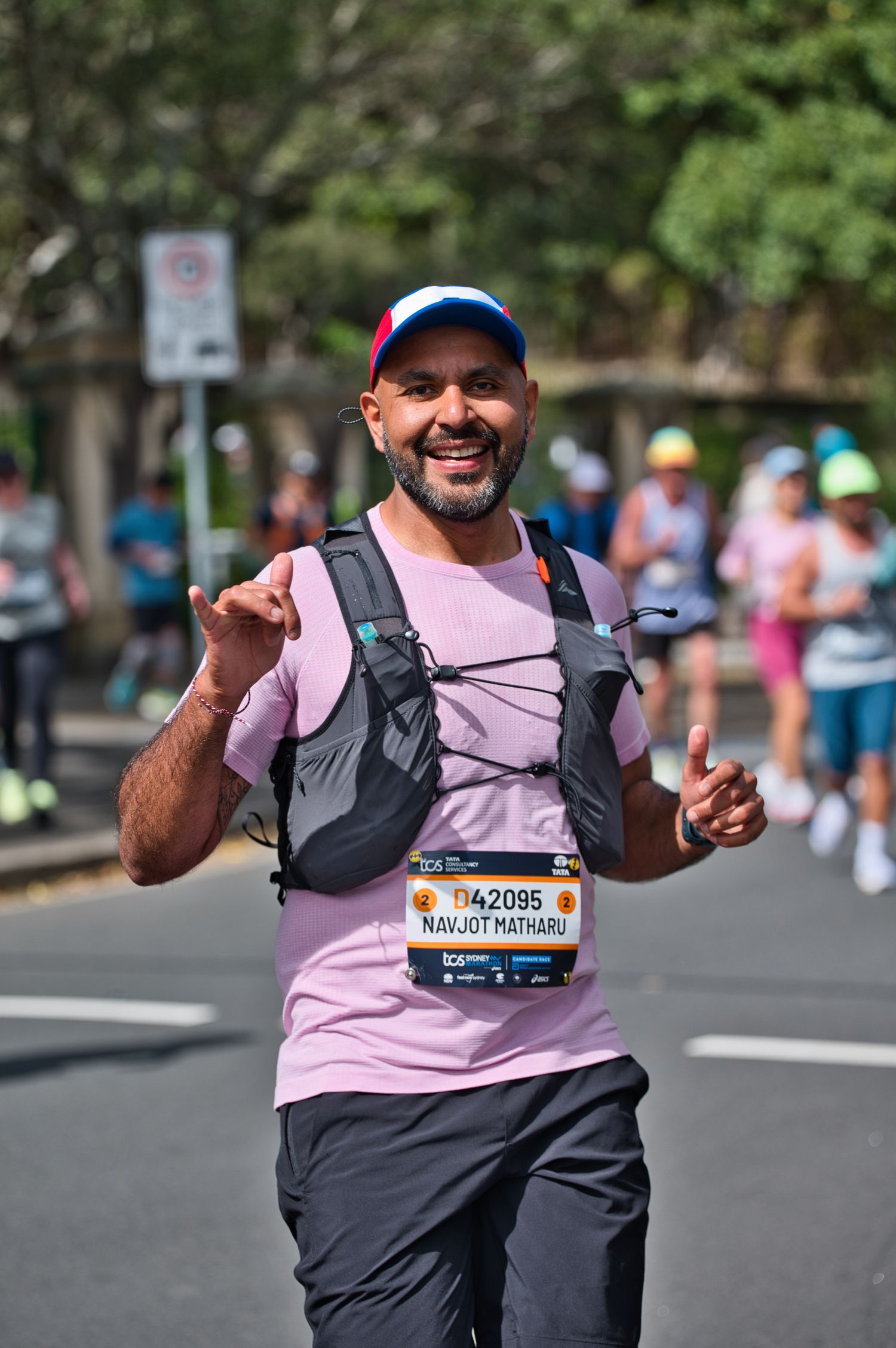 A marathon runner in pink shirt and race bib gives thumbs up while running on a city street.
