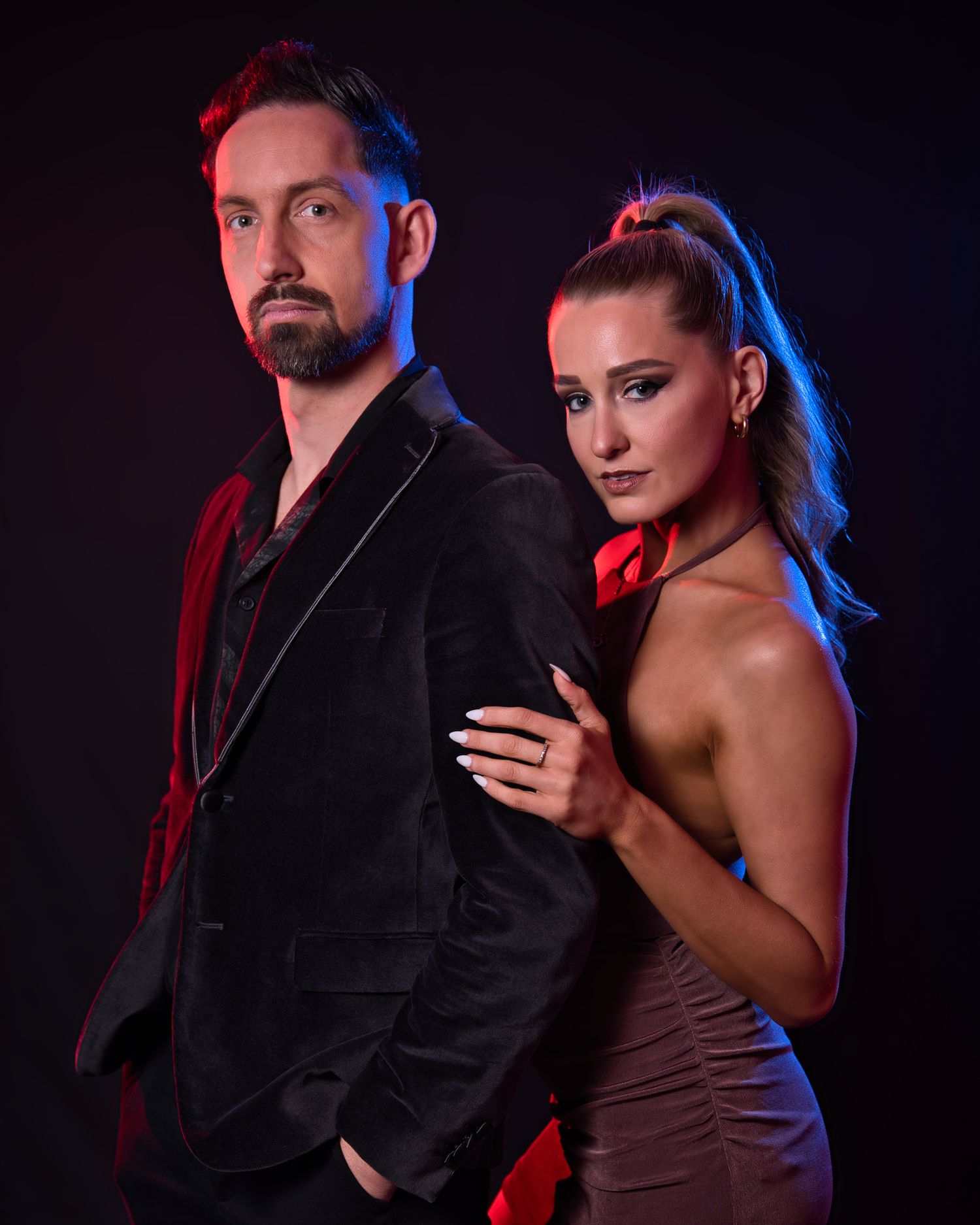 Dramatic studio portrait of dance partners in formal attire with red and blue lighting.