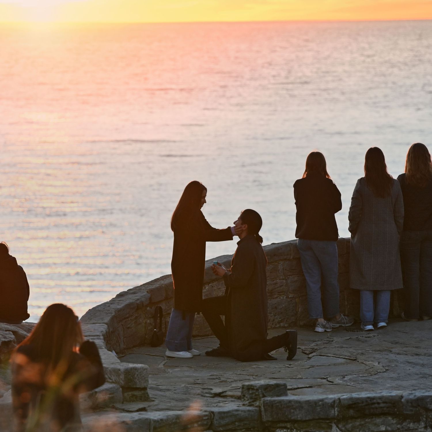 Silhouettes of people gathering on coastal rocks during golden sunset.