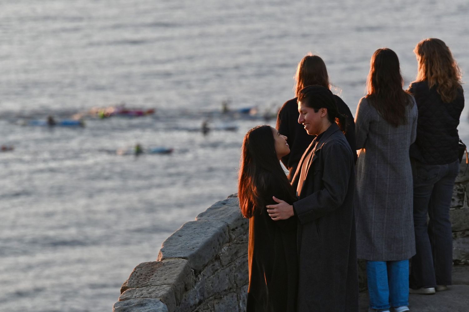 Group of onlookers at coastal viewpoint with ocean and swimmers in background.
