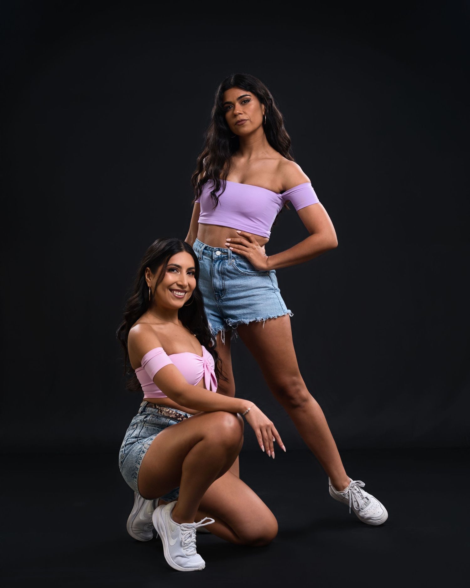 Two models pose in matching purple crop tops and denim shorts against a dark background.
