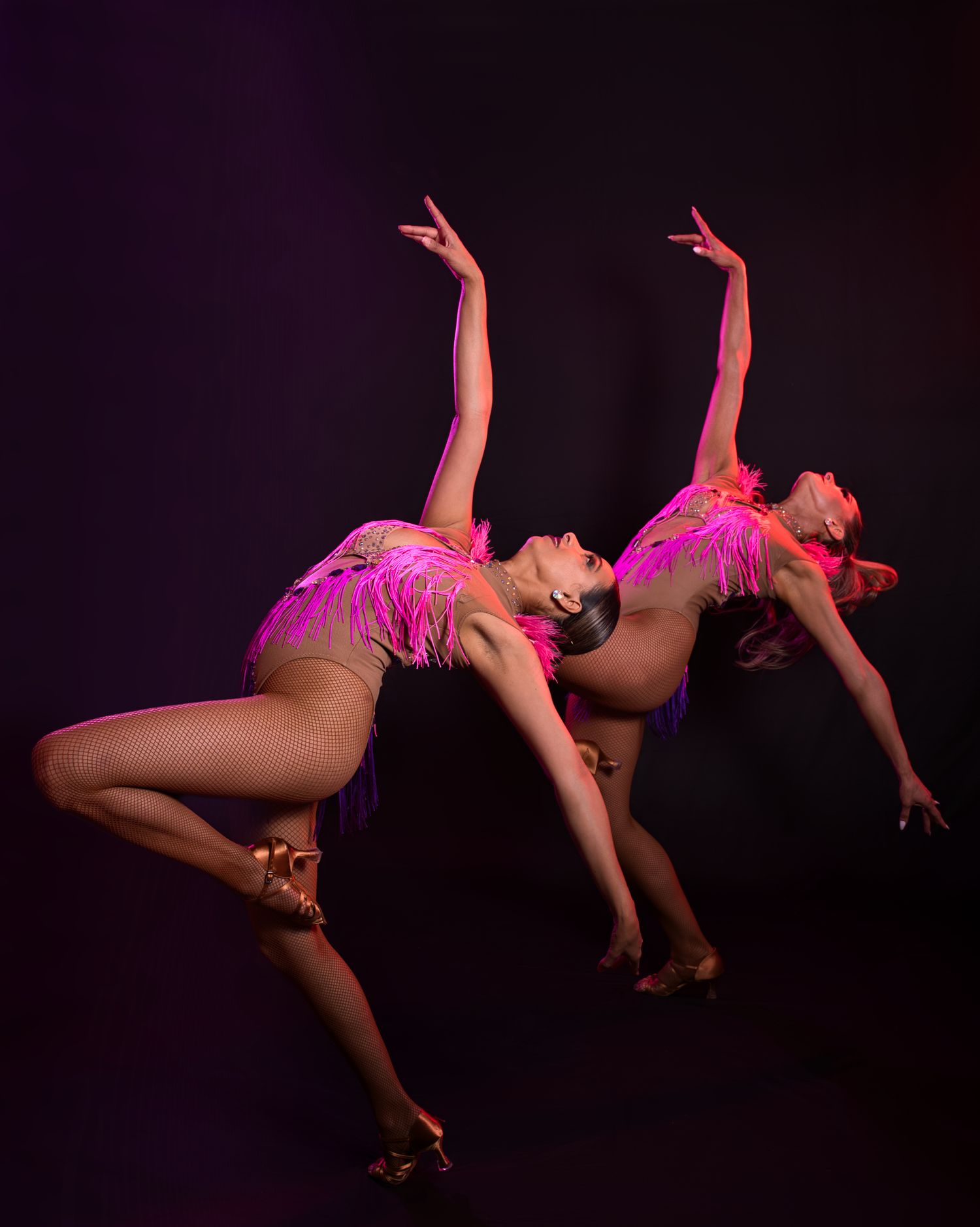 Two performers in pink costumes executing synchronized dance moves on dark stage.