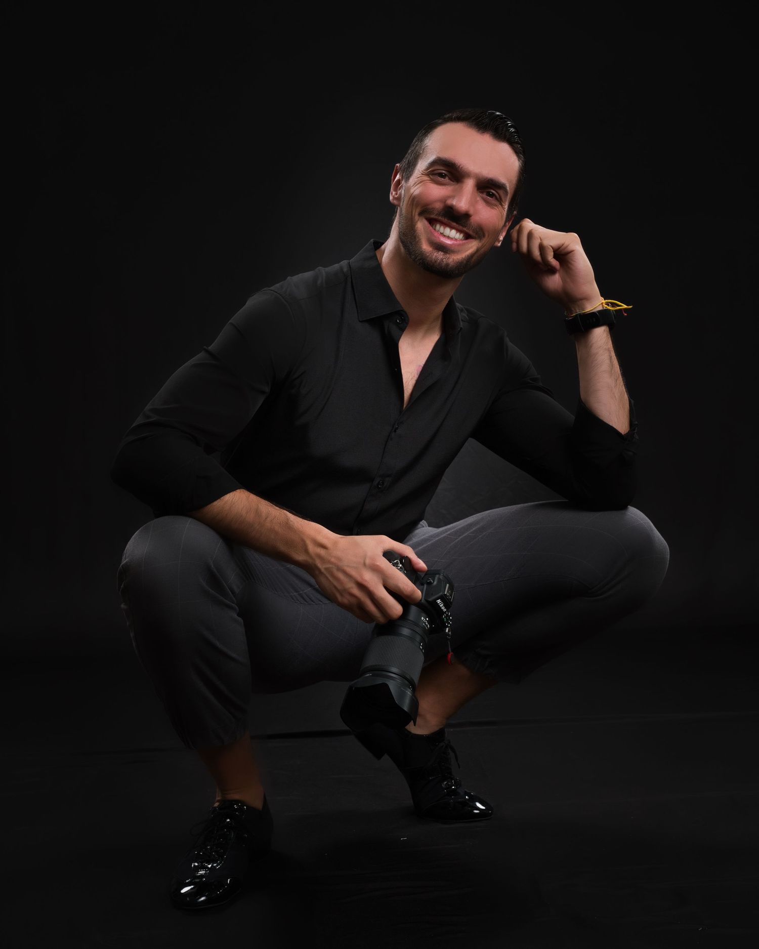 Stylish portrait of person in black outfit crouching with hand near face against dark background.