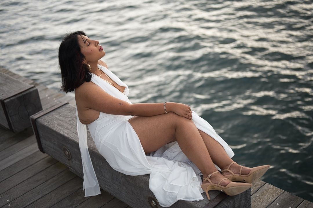 A woman in a flowing white dress relaxes on a wooden deck by the ocean.