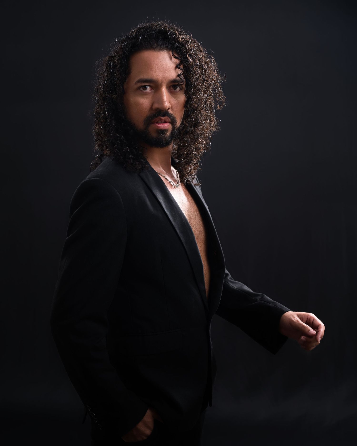 Studio portrait of a man with long curly hair wearing an open black blazer.