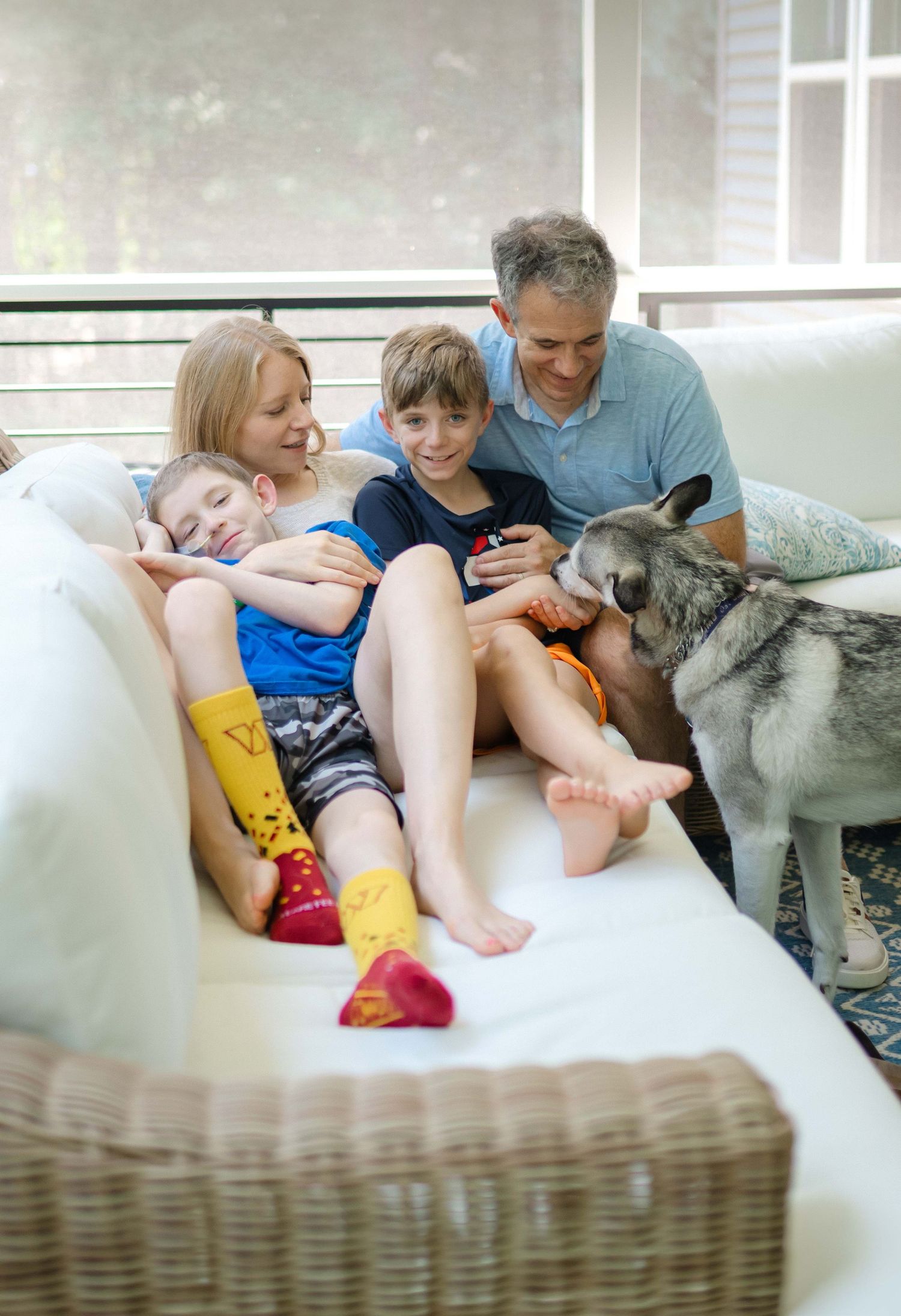 Family with two children and a husky dog relaxing together on a white wicker couch indoors.