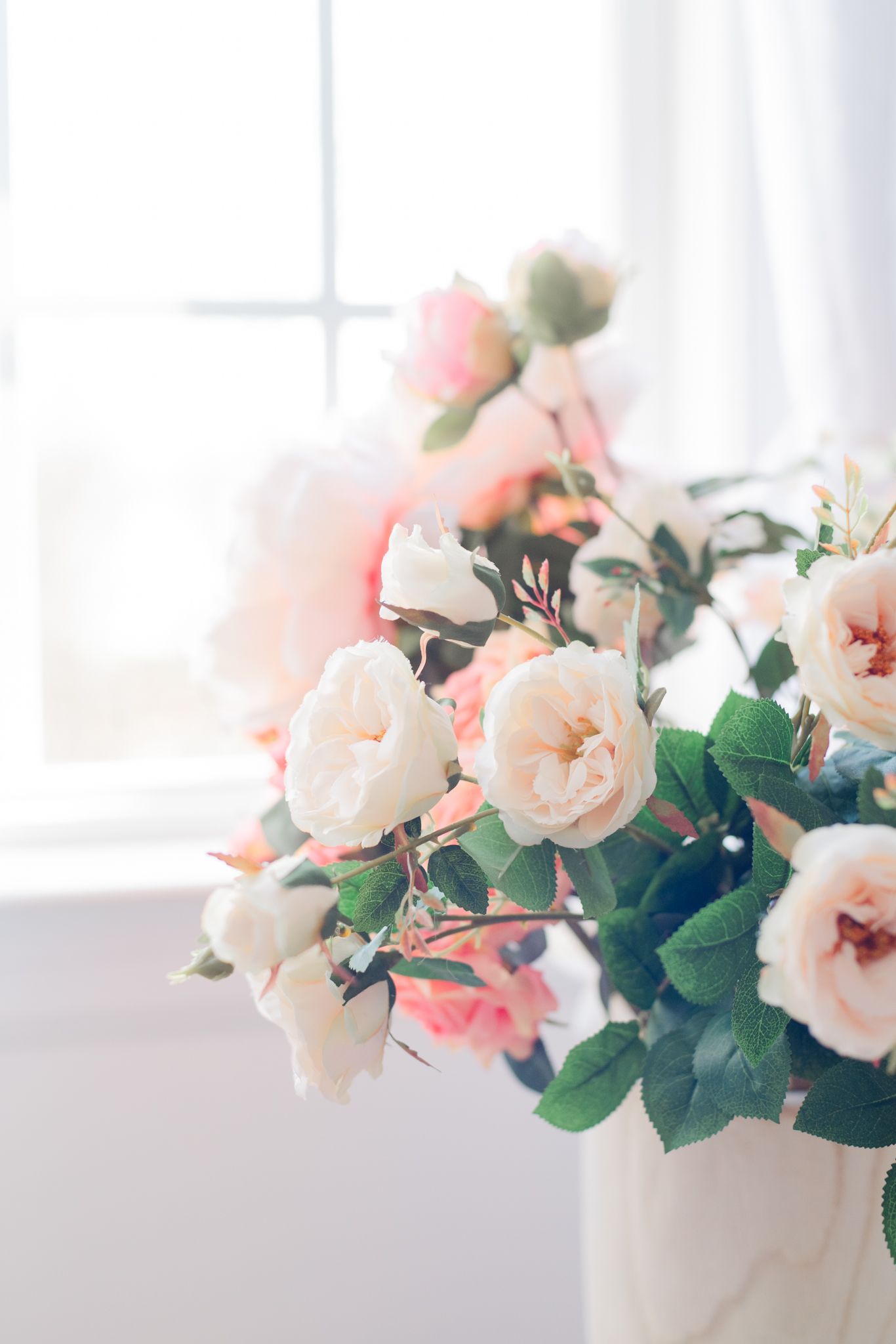 Soft pink roses arranged in white vase by sunlit window.