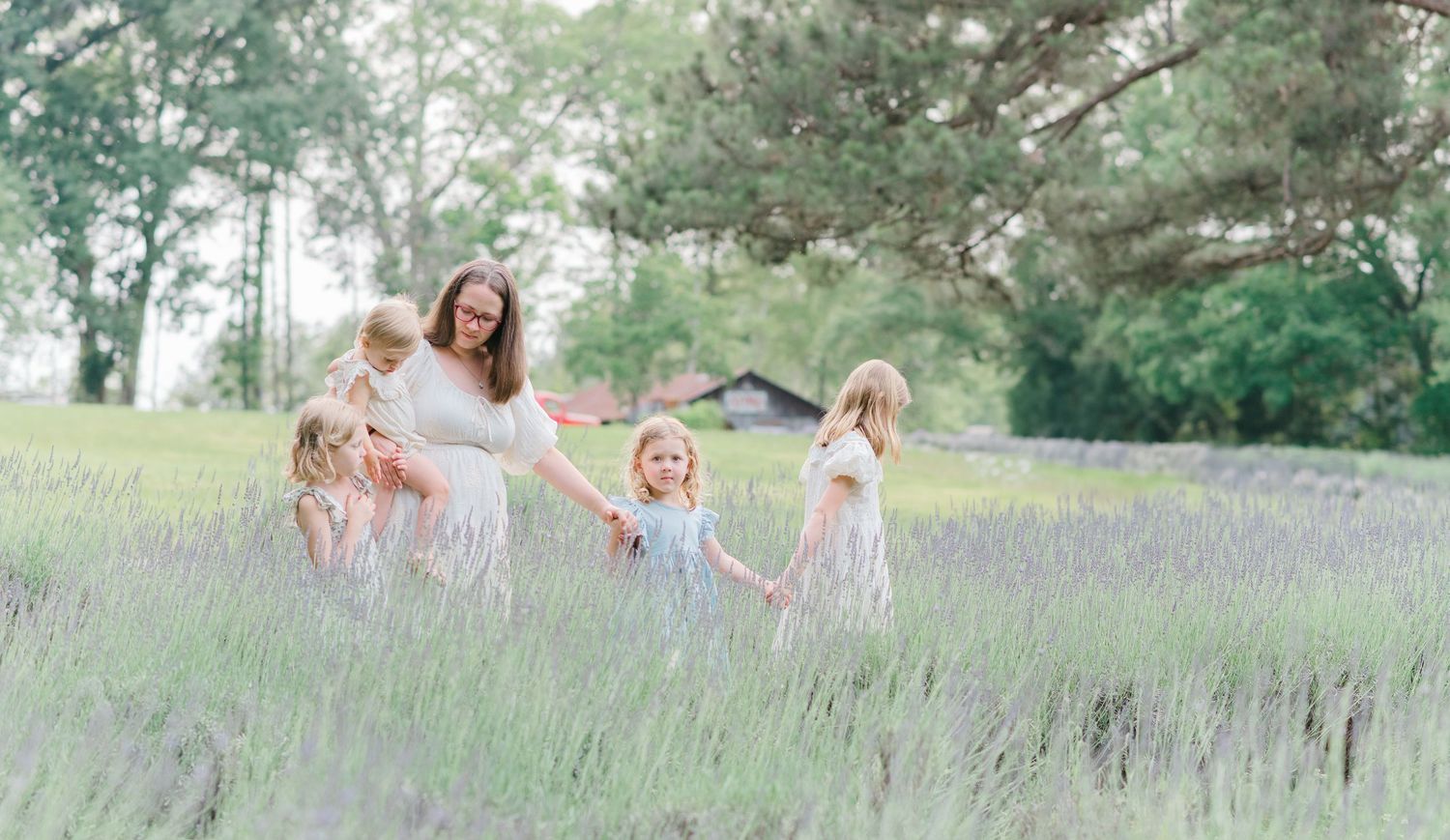 Pregnant woman in white dress surrounded by young girls holding hands in a blooming lavender field.