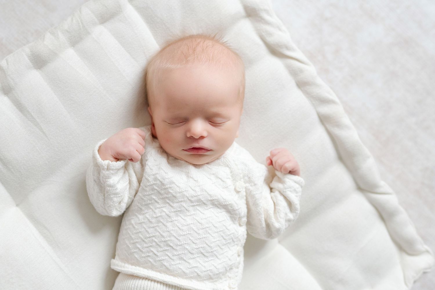 Peaceful newborn baby sleeping in white outfit on soft white blanket with arms raised.