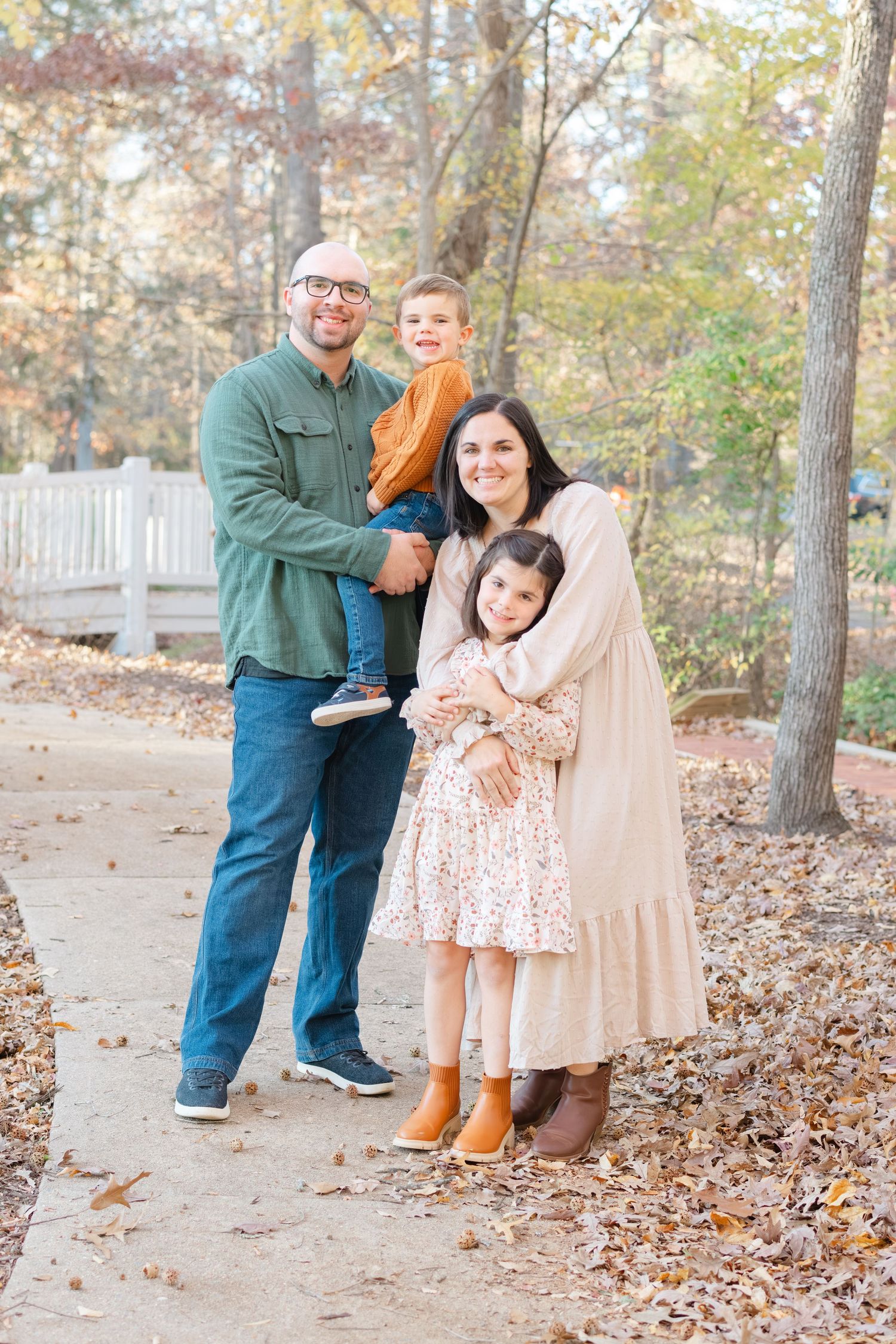 Family of four smiling outdoors in fall, parents holding two young children on a leaf-covered path among autumn trees.