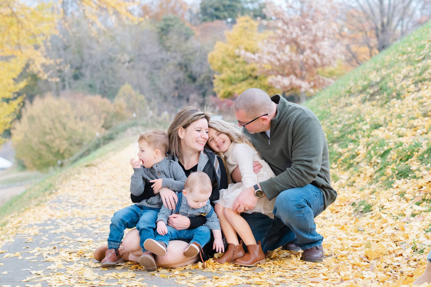Family of five sharing a warm embrace on a leaf-covered path surrounded by autumn foliage.
