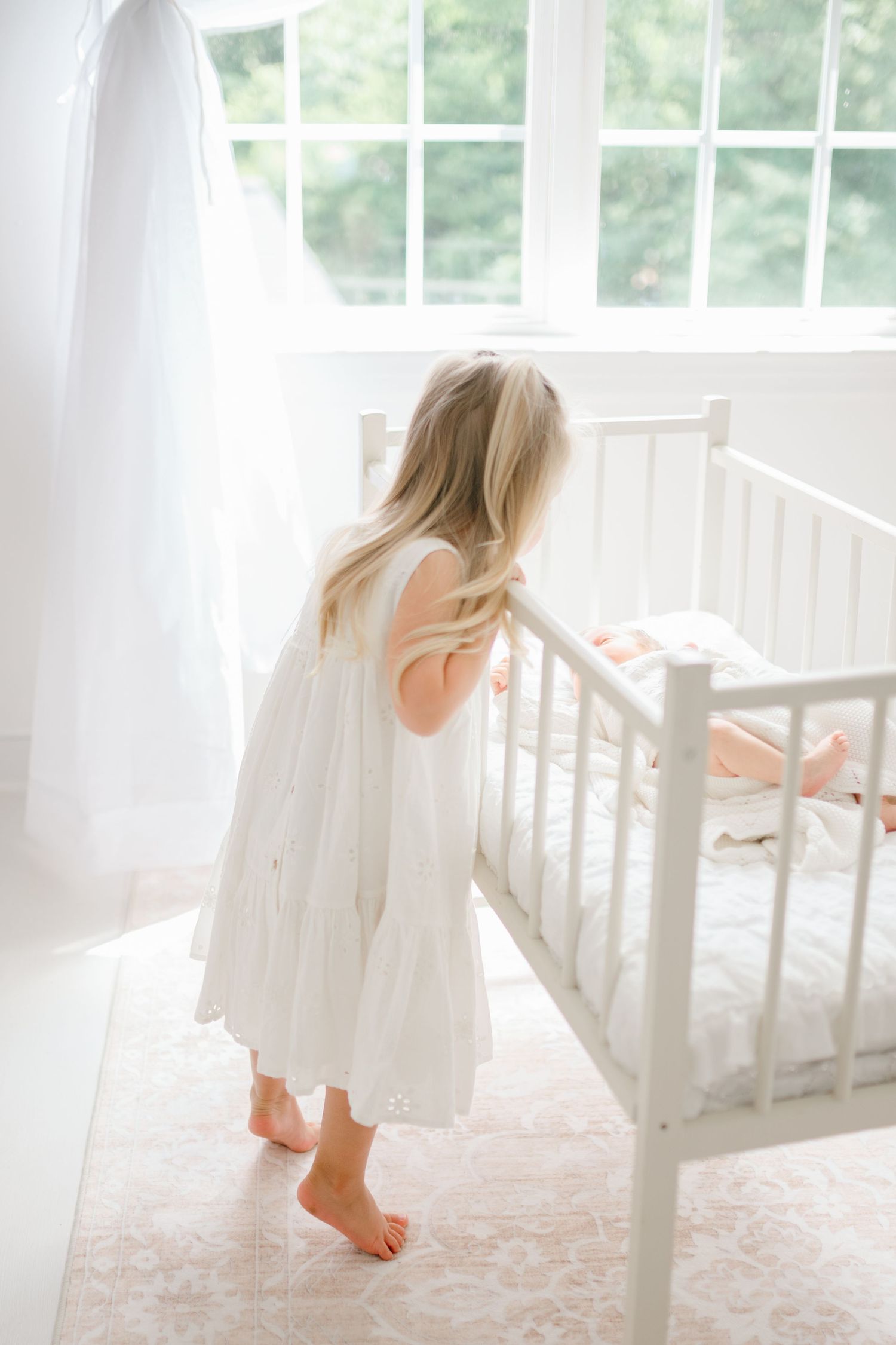 A young girl in a white dress stands barefoot beside a white crib in a bright, airy nursery room.