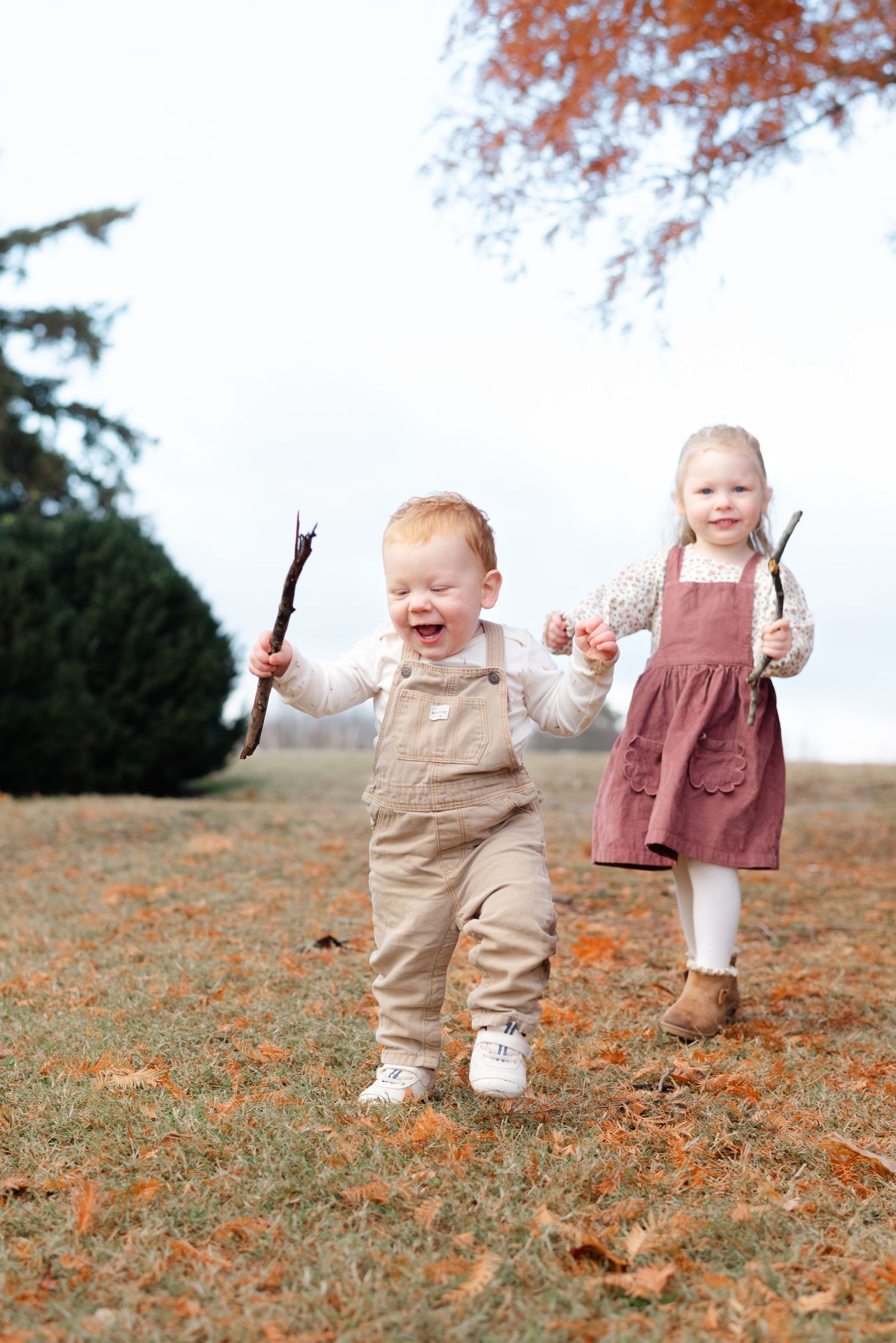 Two toddlers laughing and running outdoors in autumn, holding sticks, wearing overalls and a pinafore dress.